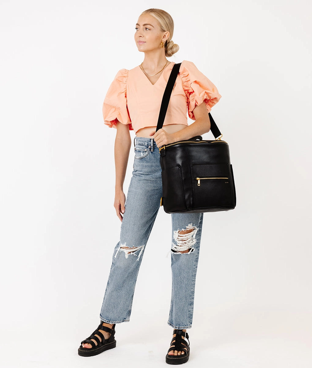 Stylish woman wearing a peach top and jeans, showcasing the Original Diaper Bag Backpack in faux leather.