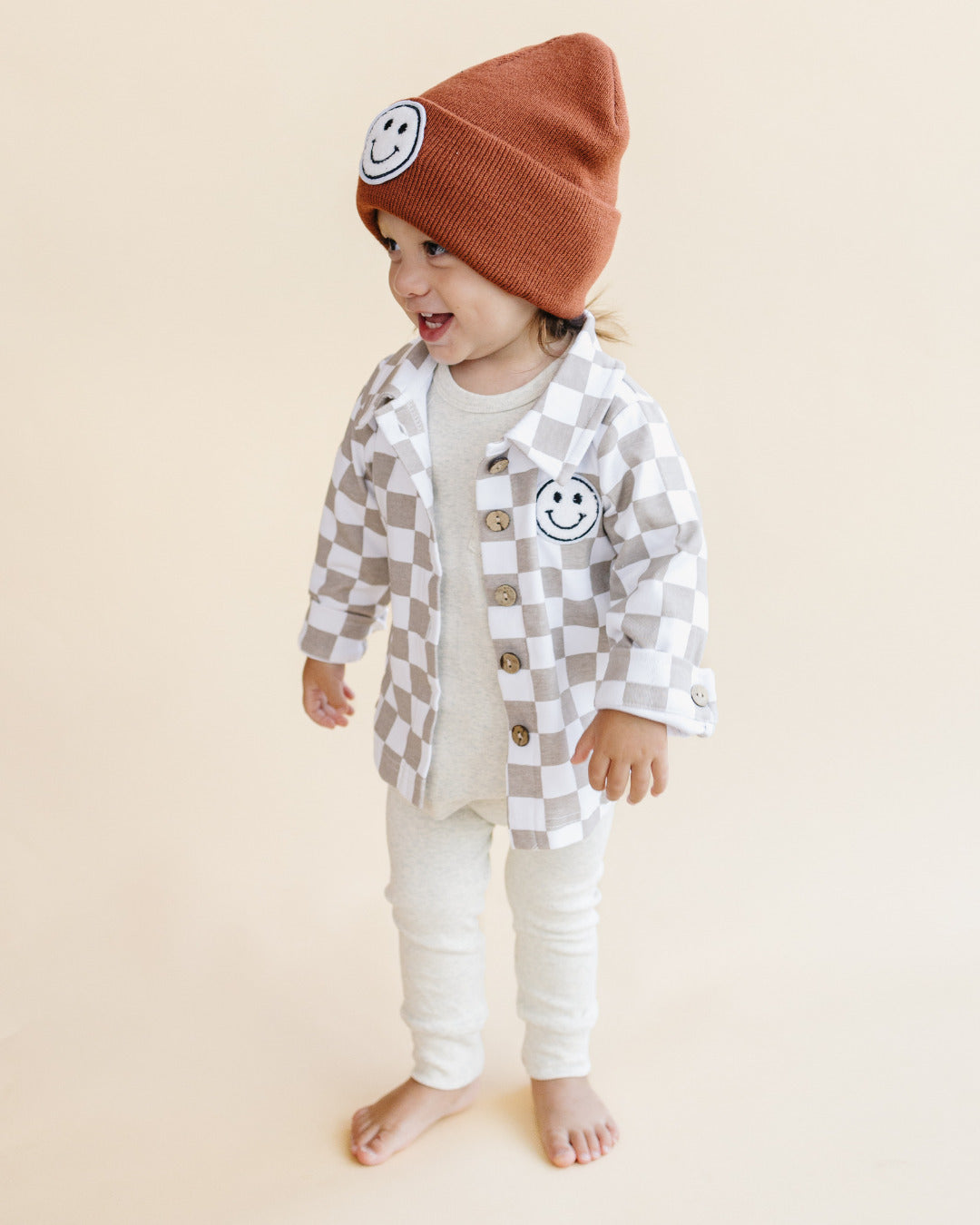 Toddler wearing Cotton Shacket in Smiley Checkered Latte with a smiley patch and brown beanie, smiling outdoors.