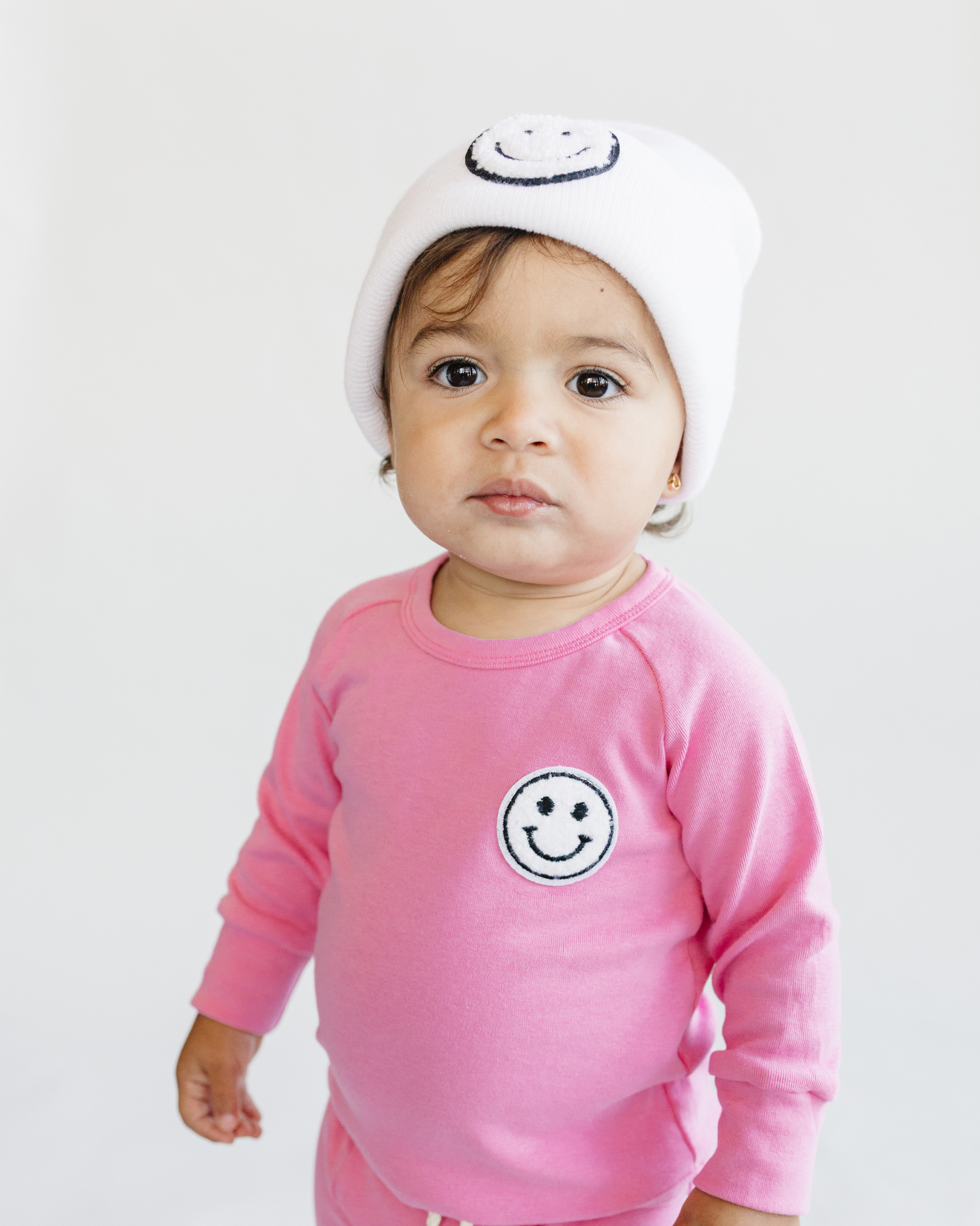 Toddler girl wearing a pink lounge set smiley bubble gum with a smiley patch and white beanie.