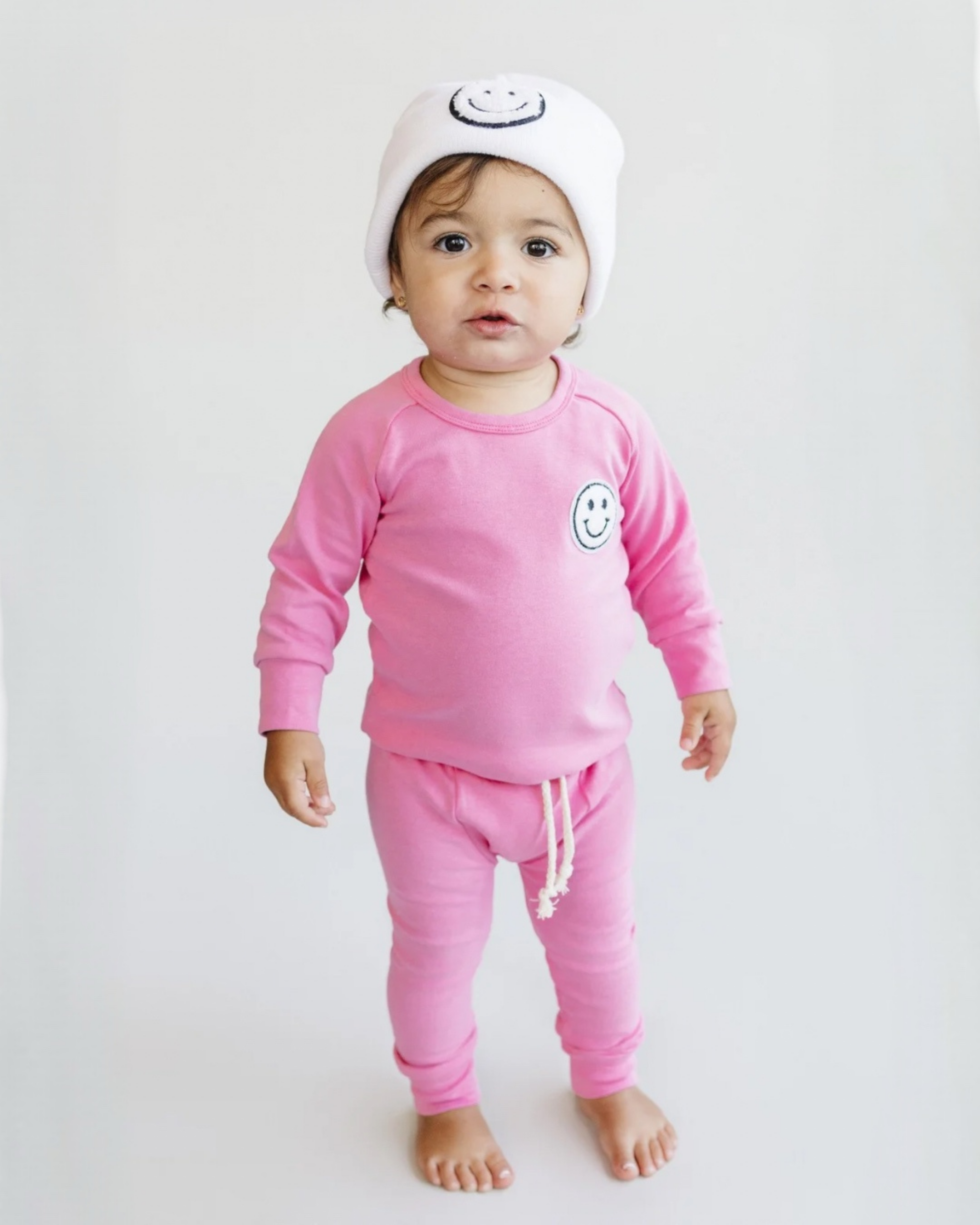 Toddler in pink lounge set smiley bubble gum with a matching white smiley beanie, showcasing comfort and style.