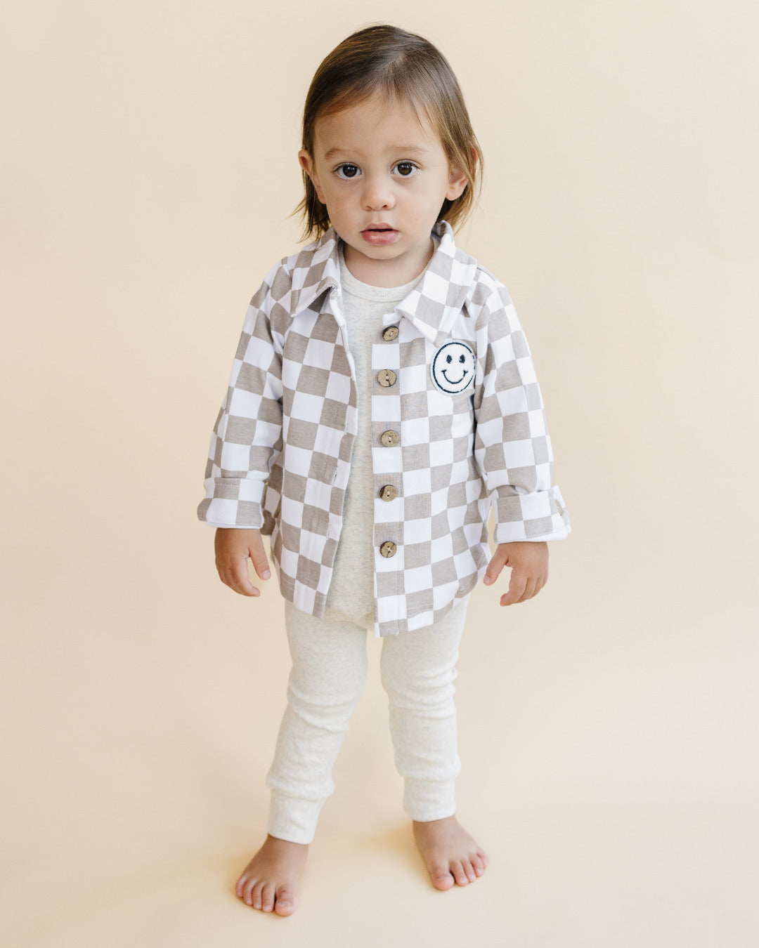 Toddler wearing a Cotton Shacket in Smiley Checkered Latte with a cheerful smiley patch and button-down design.