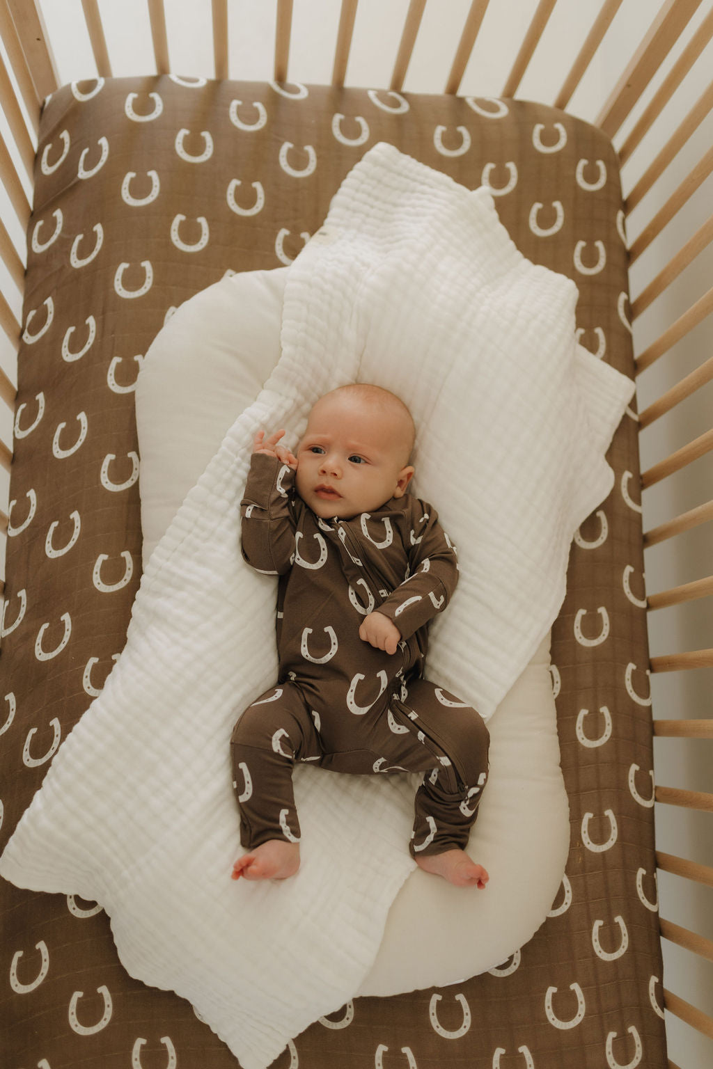 Baby wearing bamboo zip pajamas giddy up with horseshoe print in crib on cozy white blanket.