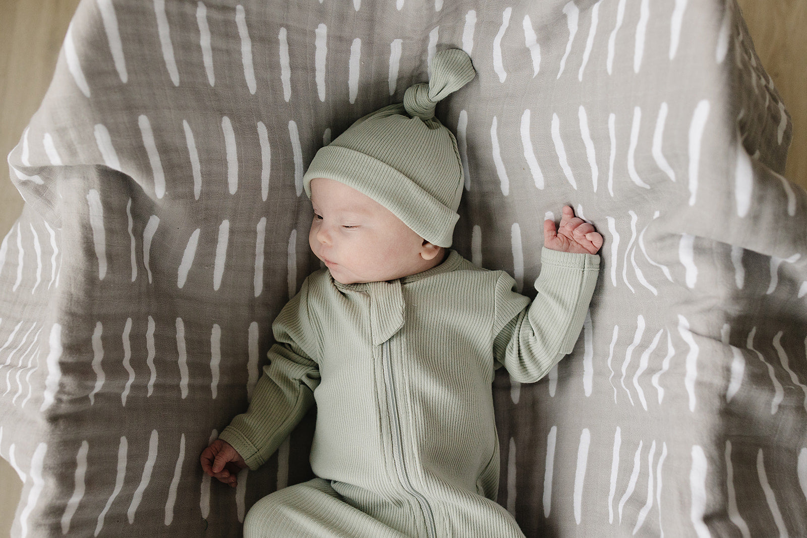 Newborn wearing a sage organic ribbed knot hat, lying on a patterned blanket, showcasing comfort and style.