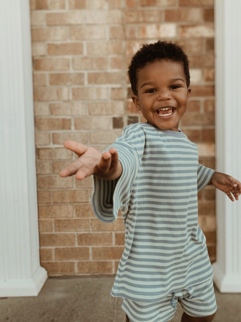 Toddler wearing a cotton candy two-piece short set, smiling and reaching out, against a brick wall background.
