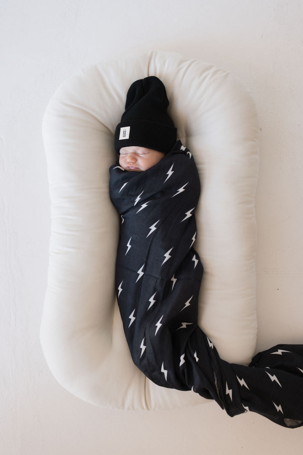 Baby wrapped in muslin swaddle midnight lightning bolt, featuring black-and-white lightning design and cozy white pillow.
