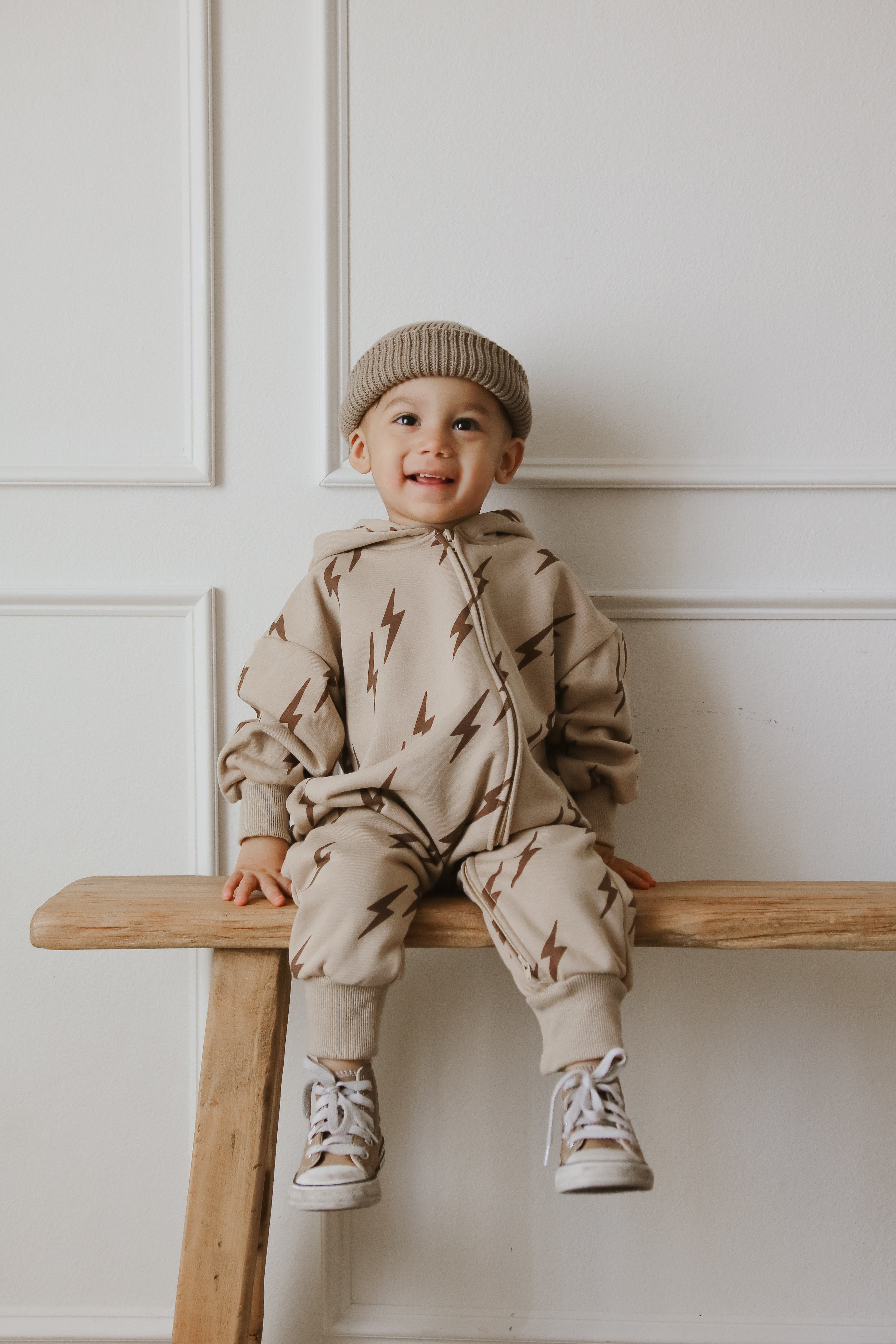 Toddler wearing a beige zip jumpsuit with lightning bolts, sitting on a wooden bench, smiling.