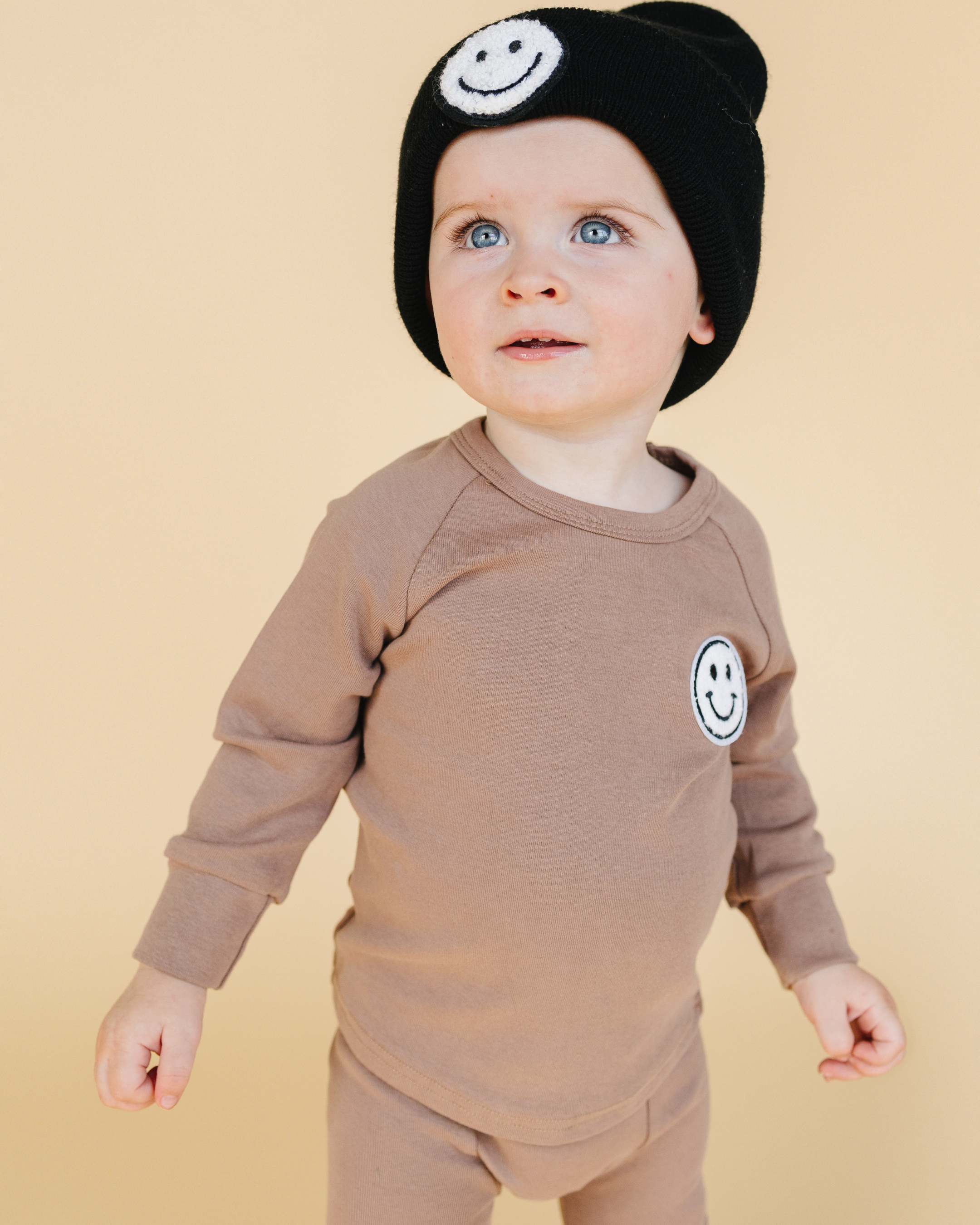 Cute toddler in lounge set smiley mocha with a black beanie featuring a smiley patch, showcasing cozy and stylish comfort.