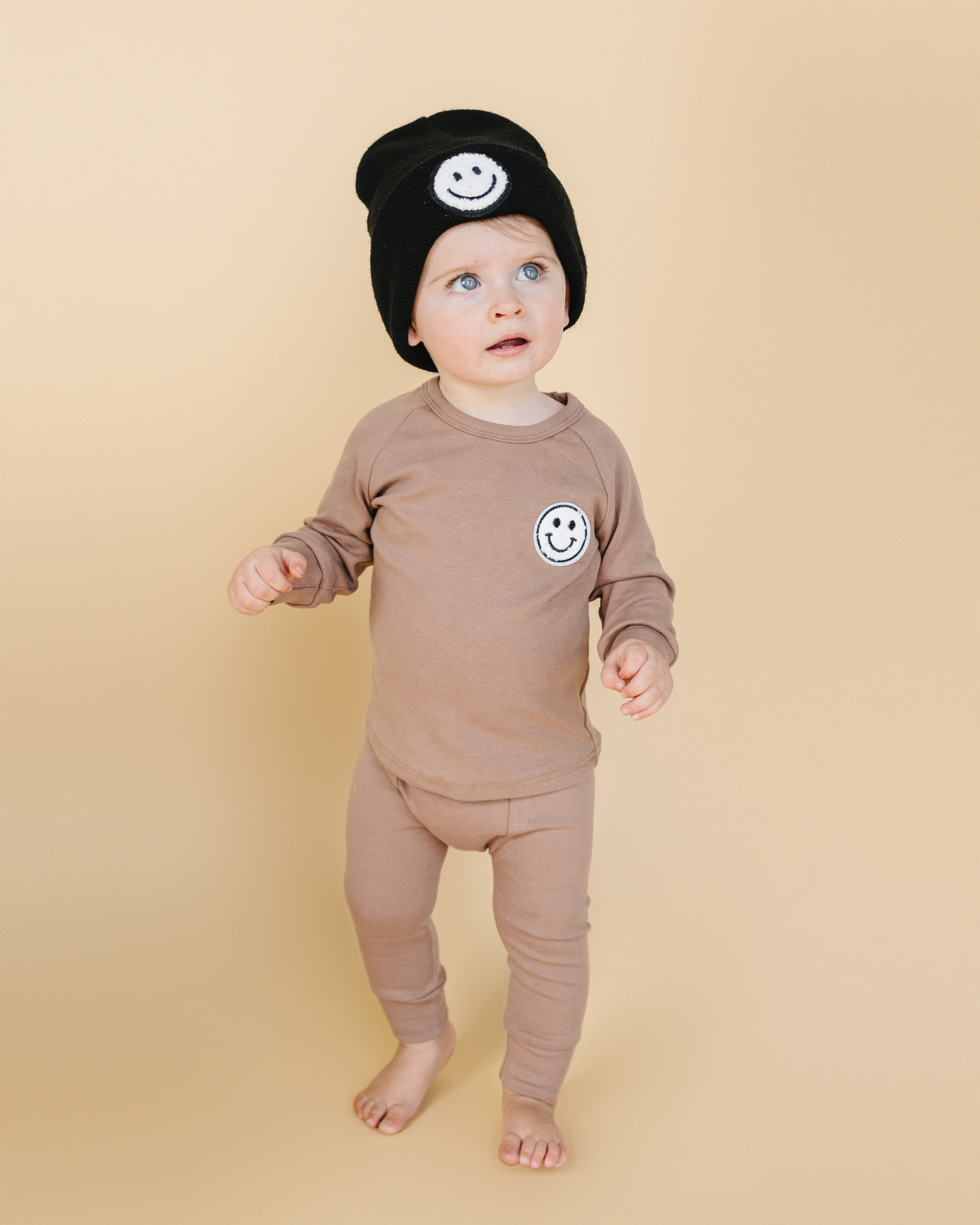 Toddler wearing Lounge Set Smiley Mocha with smiley patch and black beanie against a neutral background.