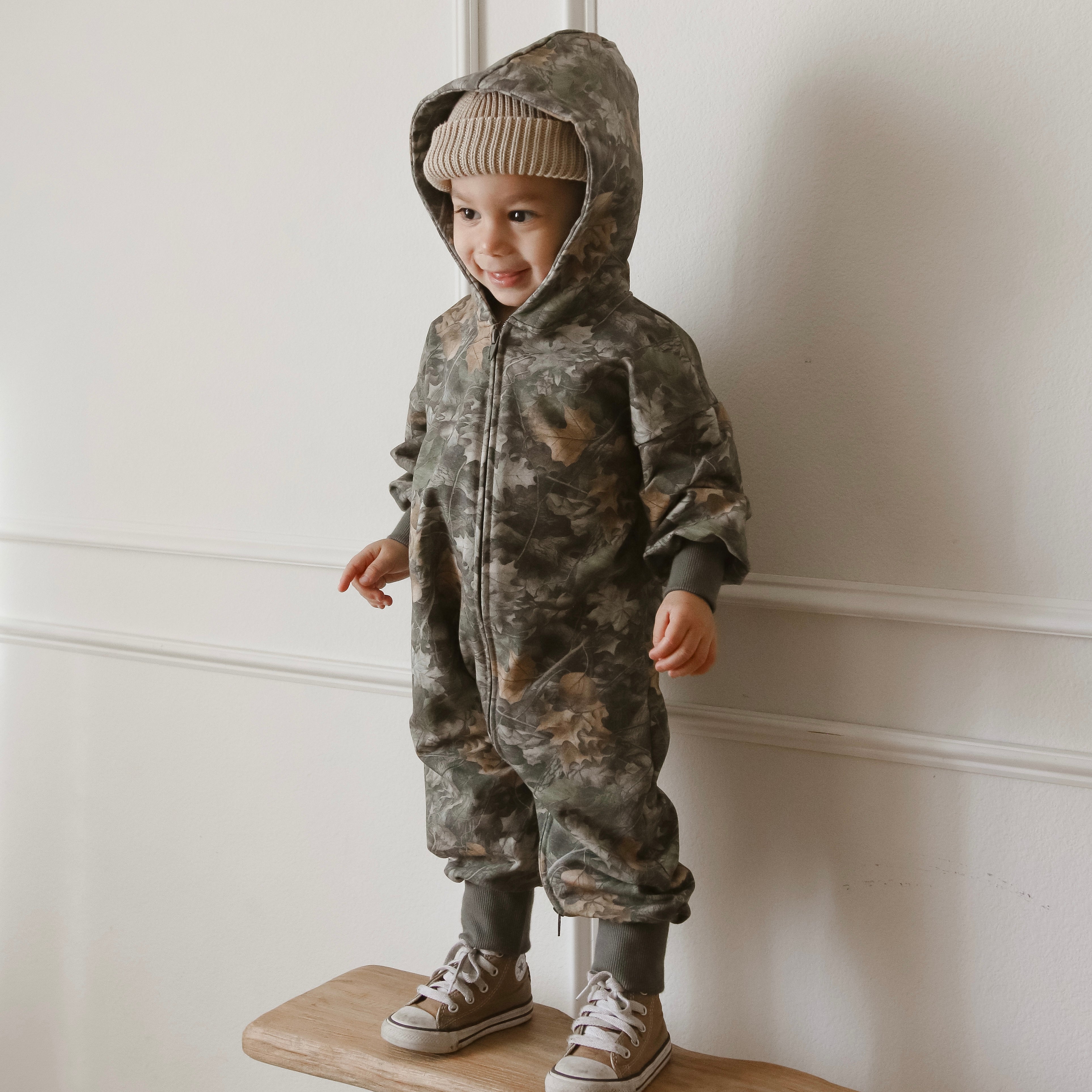 Baby wearing camo zip jumpsuit with hood, smiling on a wooden platform in a minimalist room.