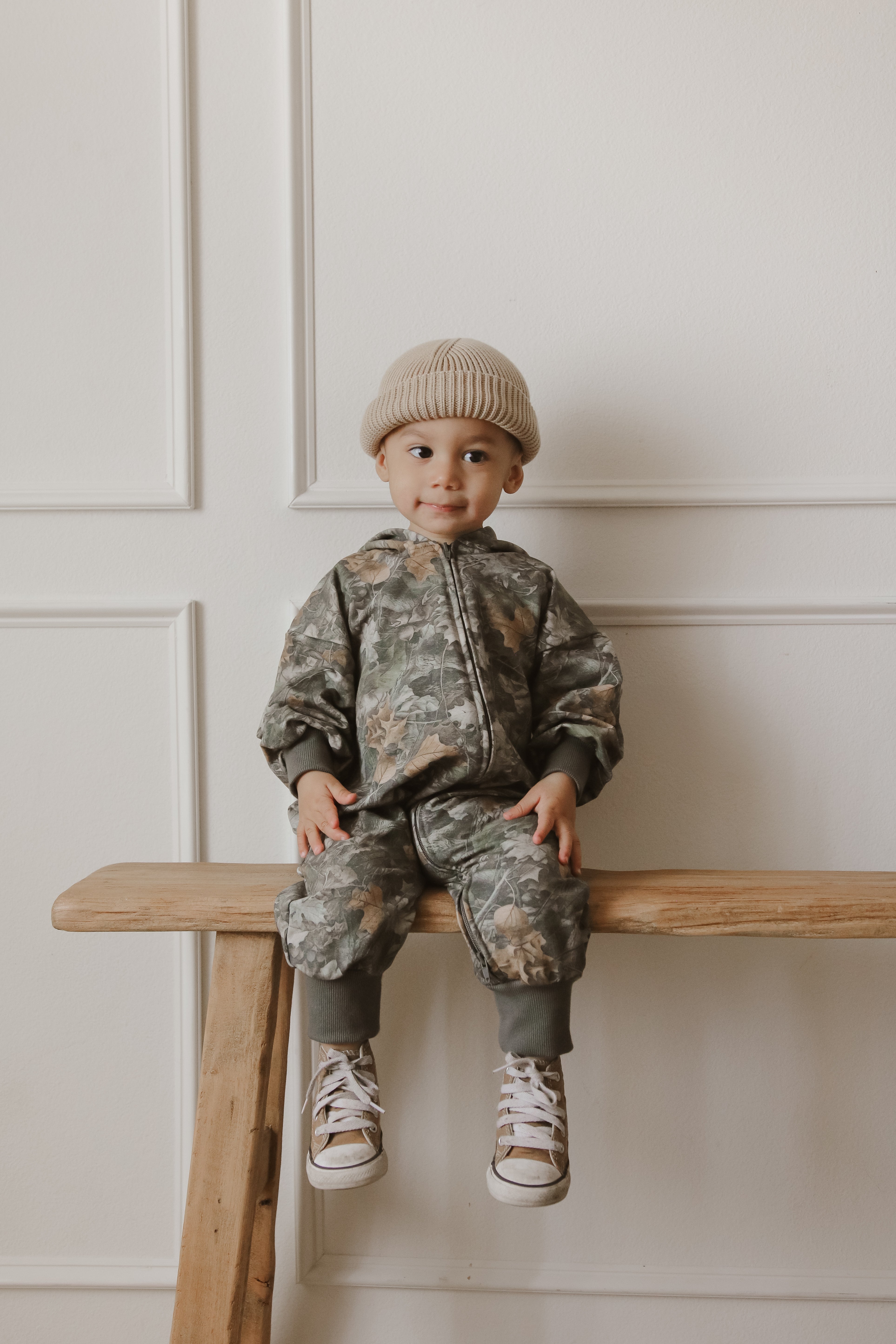 Baby in camo zip jumpsuit with beanie sitting on a wooden bench, showcasing stylish kids' clothing.
