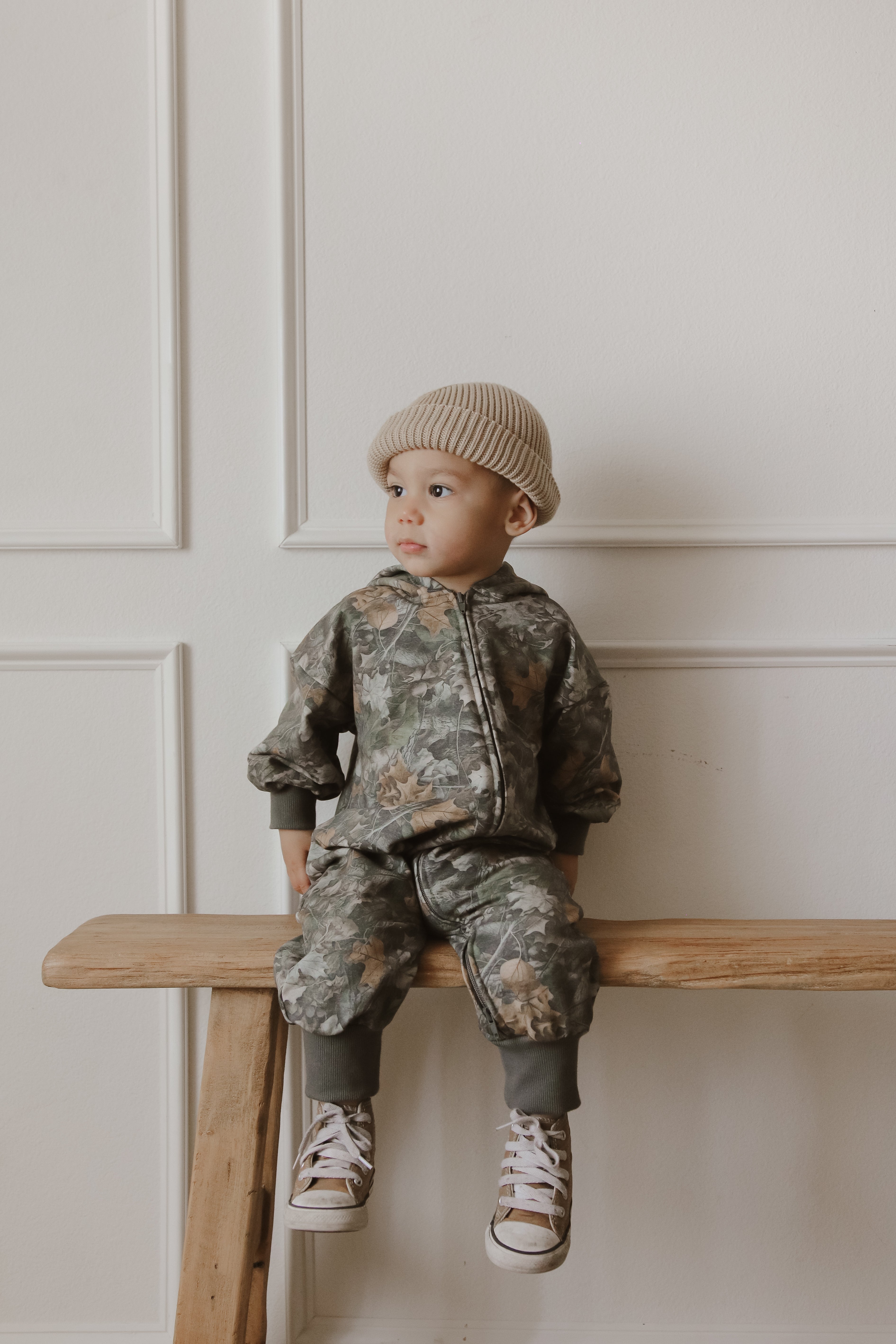 Baby wearing a camo zip jumpsuit, sitting on a wooden bench with a neutral background.