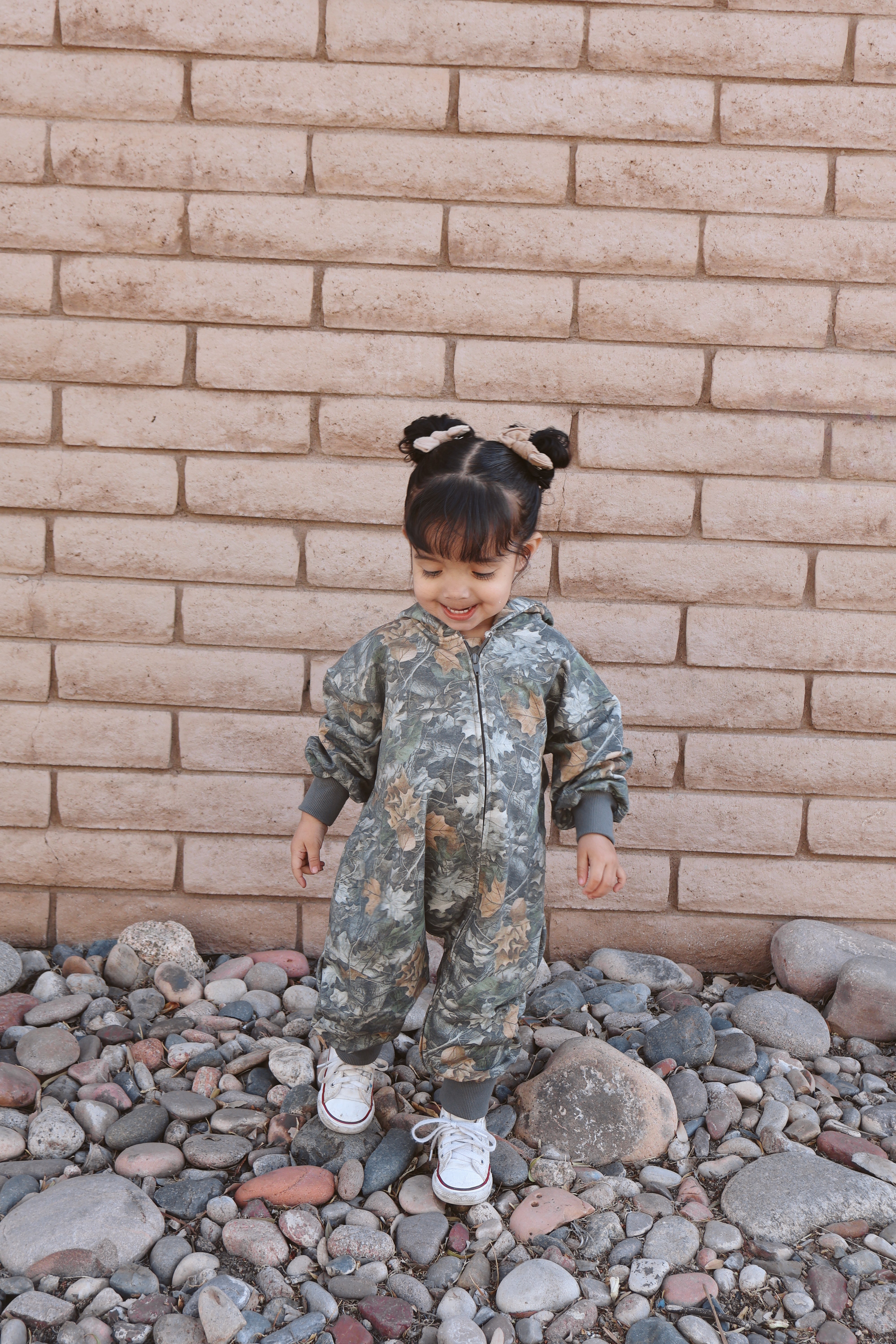 Toddler wearing a camo zip jumpsuit, standing on pebbles against a textured wall.