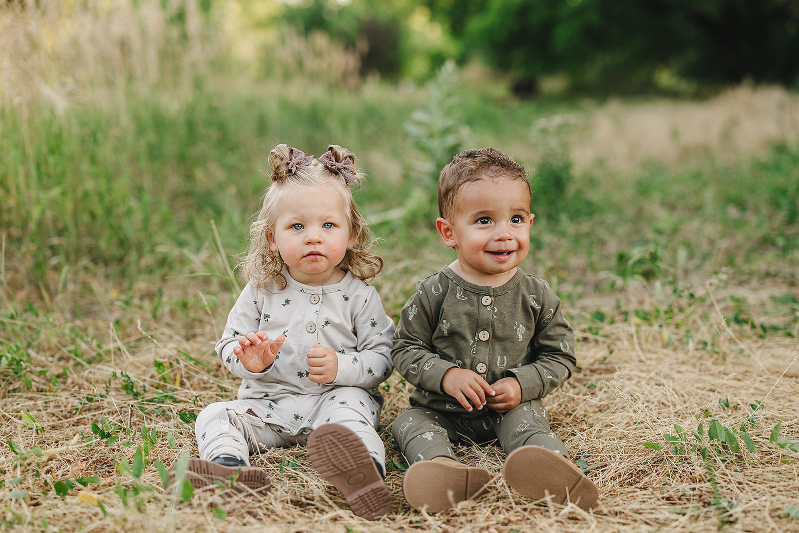 Two toddlers in cozy button rompers sitting on grass, surrounded by nature, showcasing soft styles for young children.