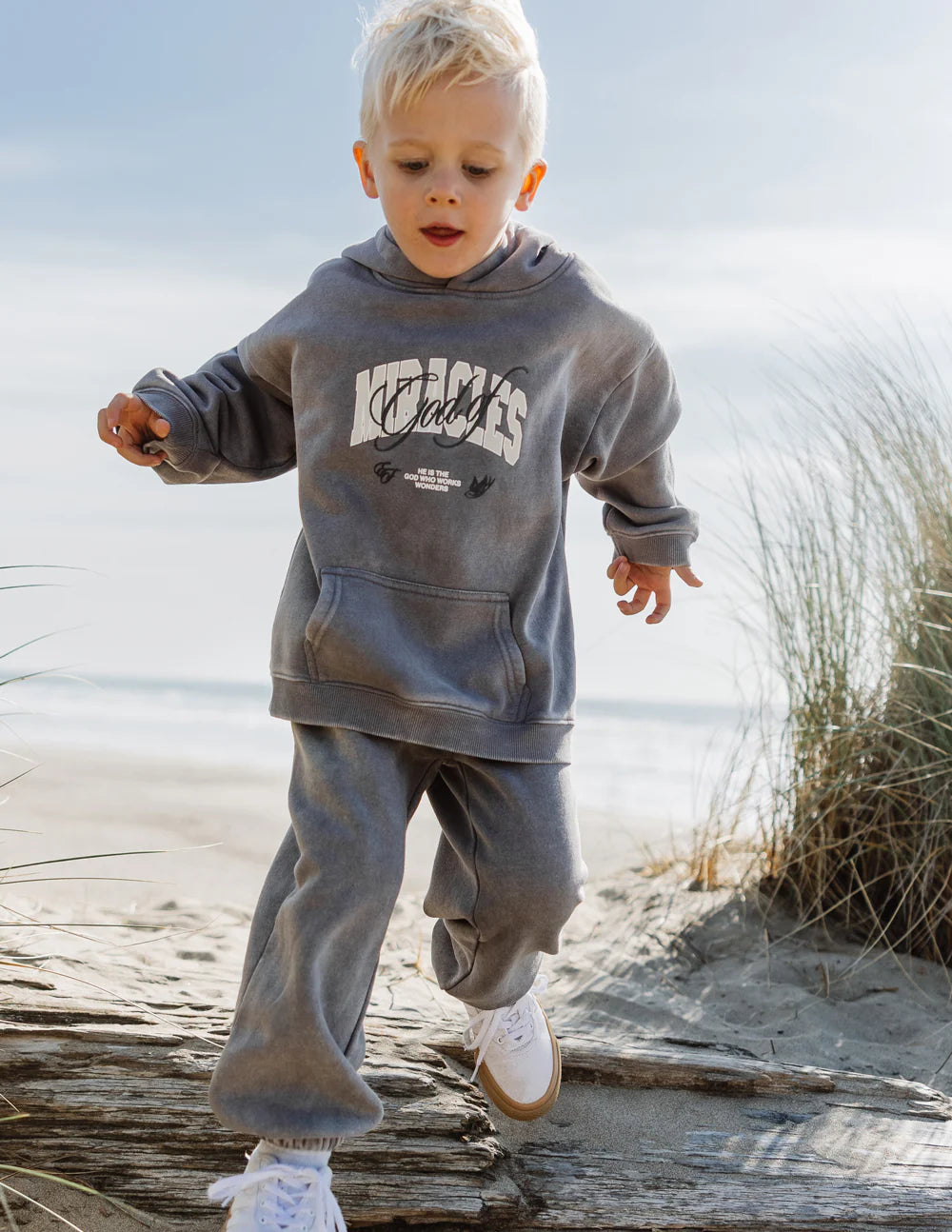 Boy wearing a gray God of Miracles kids hoodie at the beach, showcases comfort and style for kids.
