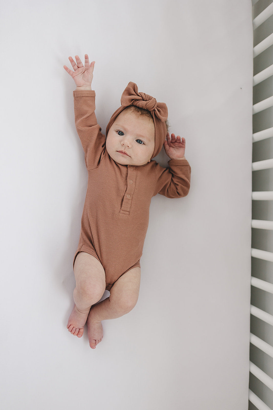 Infant wearing a brown organic baby bodysuit with a matching bow headband, resting on a crib.