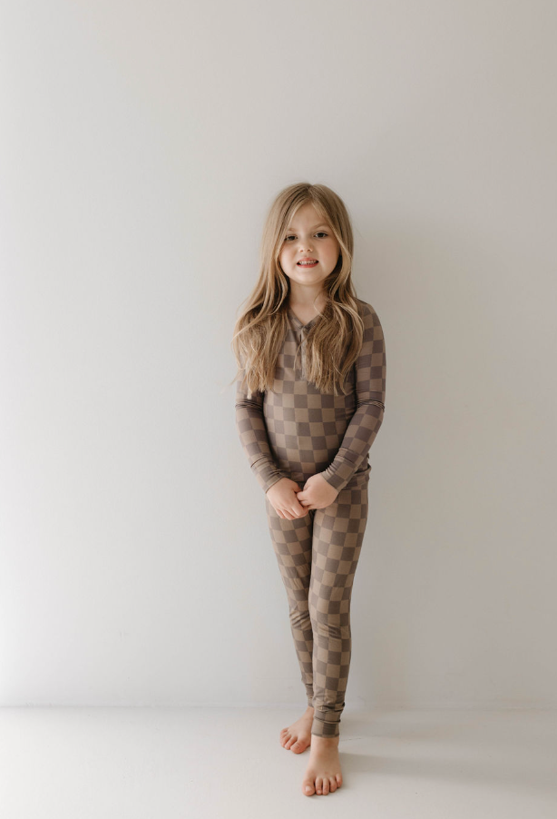 Cute girl wearing faded brown checkerboard bamboo pajamas, standing in a bright room with a smile.