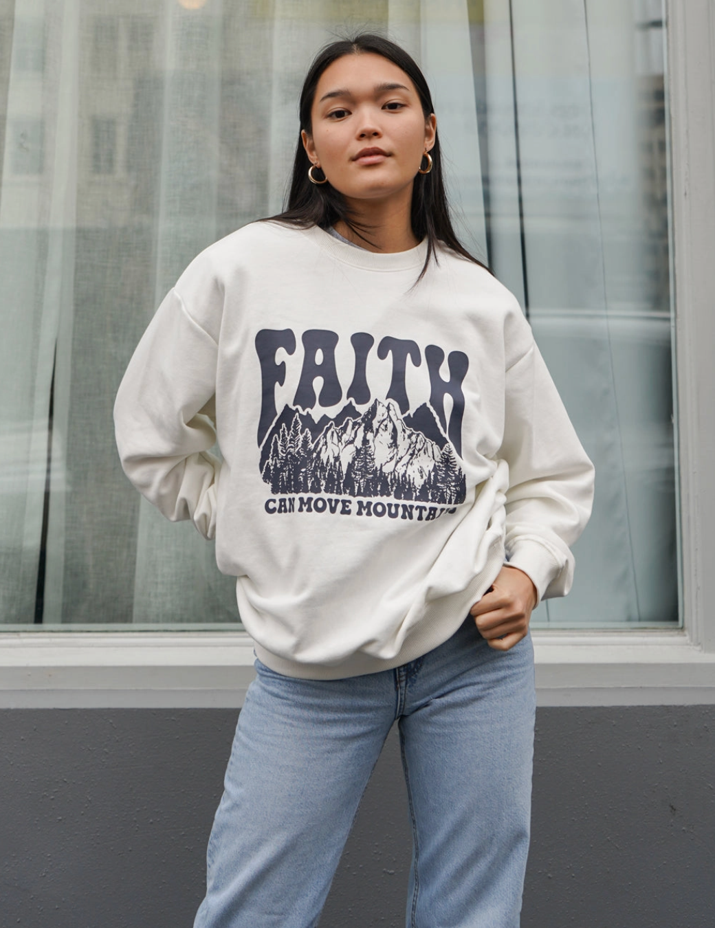Woman wearing Faith Can Move Mountains crewneck sweatshirt with mountain design and bold lettering.