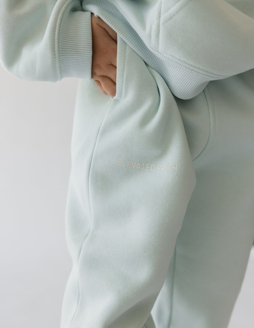 Close-up of a kid's leg in light blue sweatpants featuring a pocket and 'Elevated Death' embroidery.