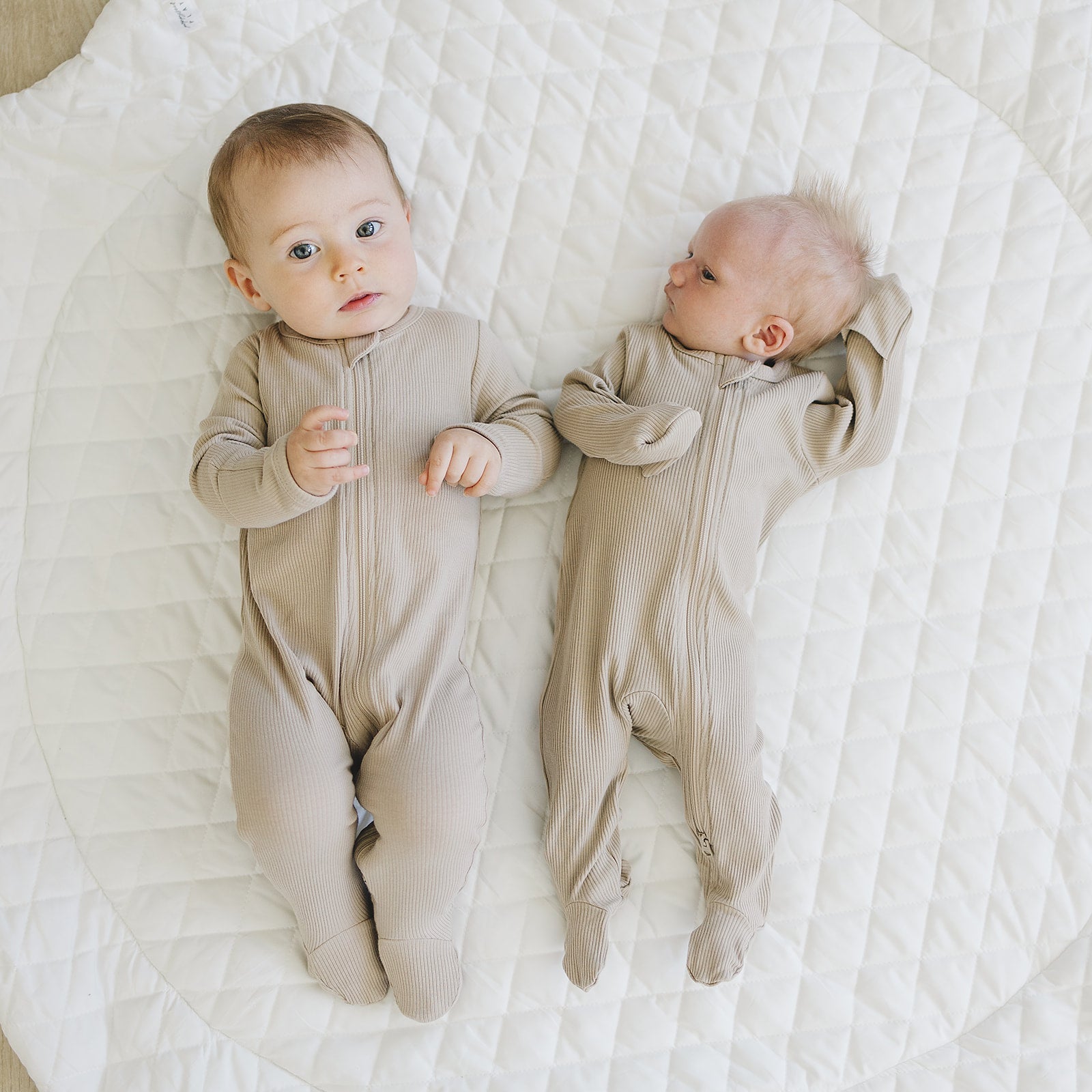 Two babies in organic baby zipper pajamas, oatmeal ribbed cotton, laying on a quilted blanket.