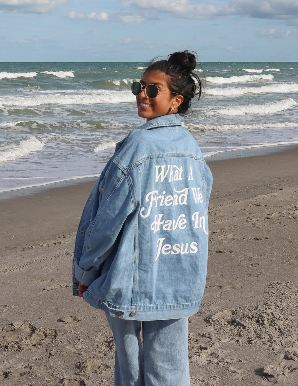 Woman wearing What a Friend in Jesus Denim Jacket by the beach, showcasing the faith-inspired message on the back.