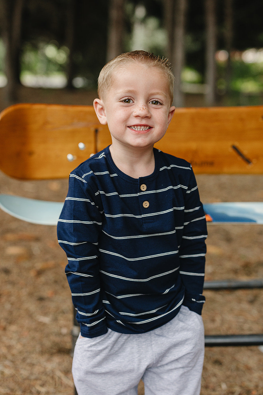 Smiling boy wearing a navy stripe long sleeve tee for kids, standing by a park bench.