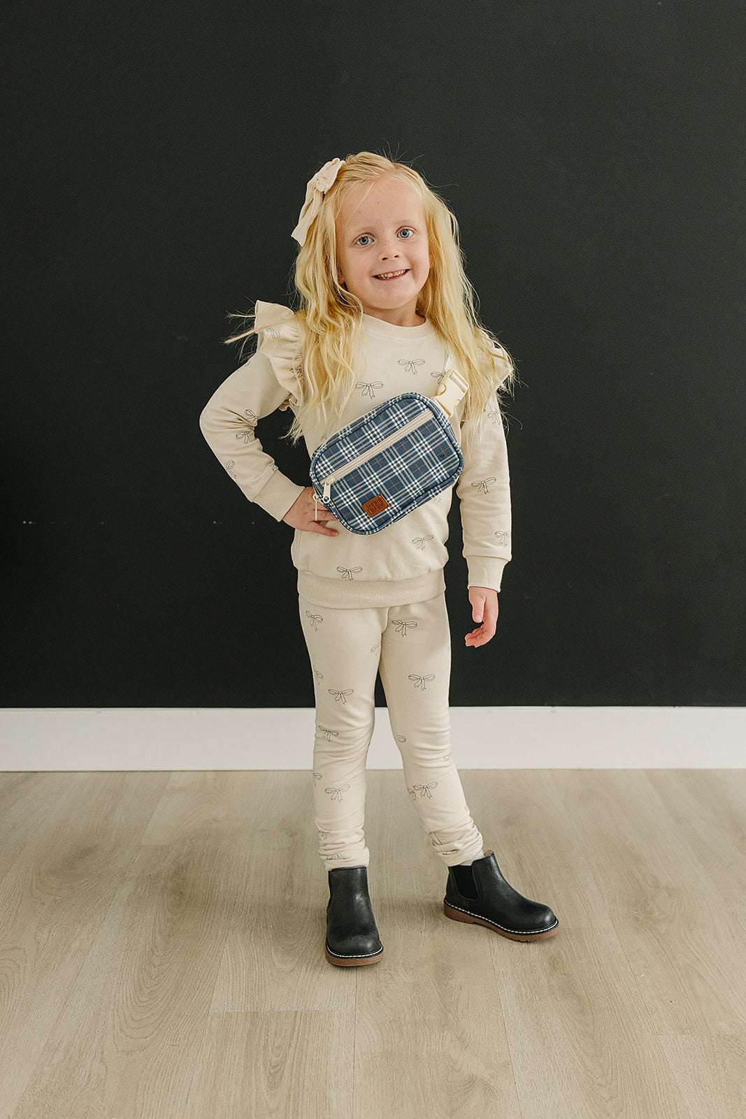A child wearing a cream outfit and a navy plaid mini fanny pack, smiling against a dark background.