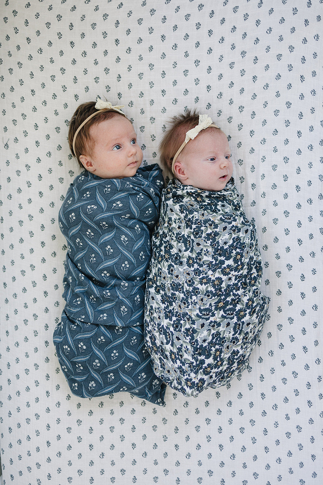 Two babies wrapped in Muslin Swaddle Blankets – one in a blue pattern and one in Liberty Floral design, on a patterned surface.