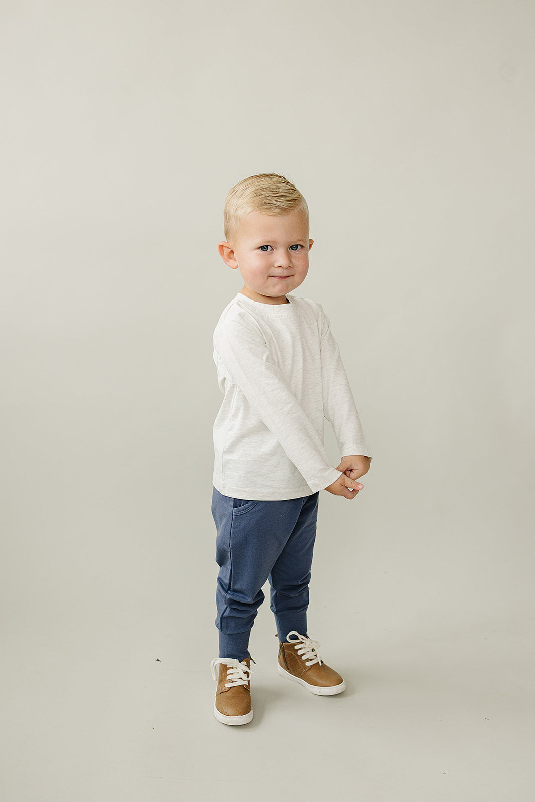 Baby wearing navy kids jogger pants, white long-sleeve shirt, and brown shoes, smiling and posing.