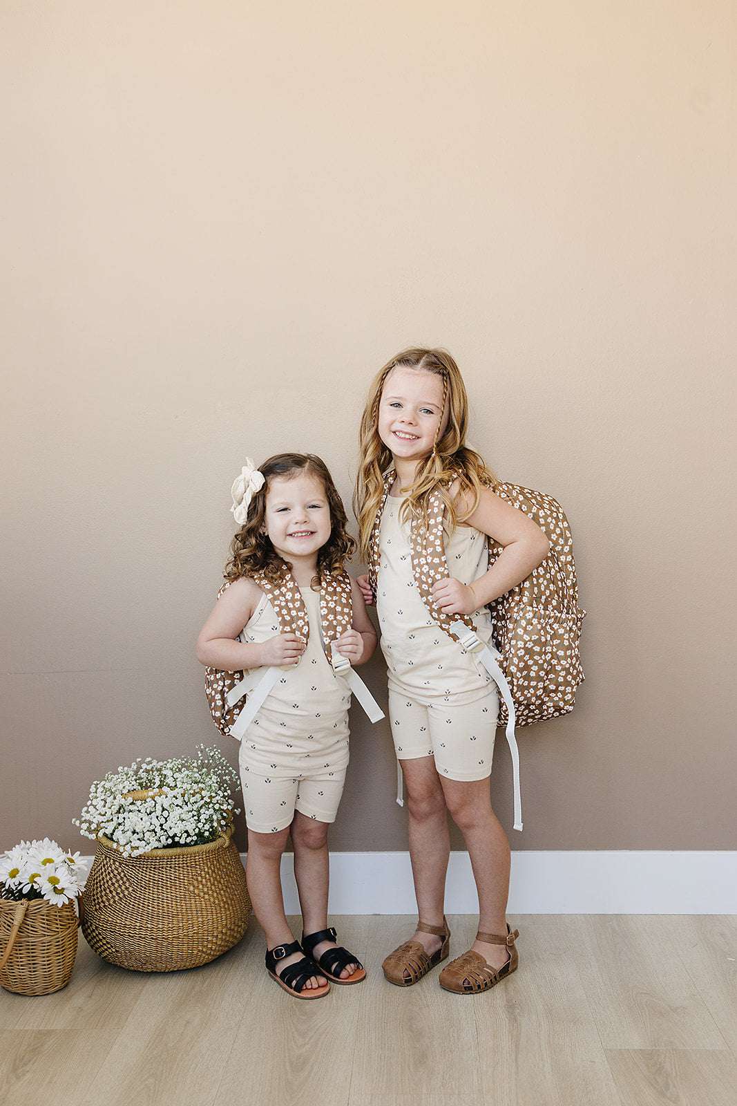 Two young girls wearing beige outfits and carrying floral backpacks, ready for school.