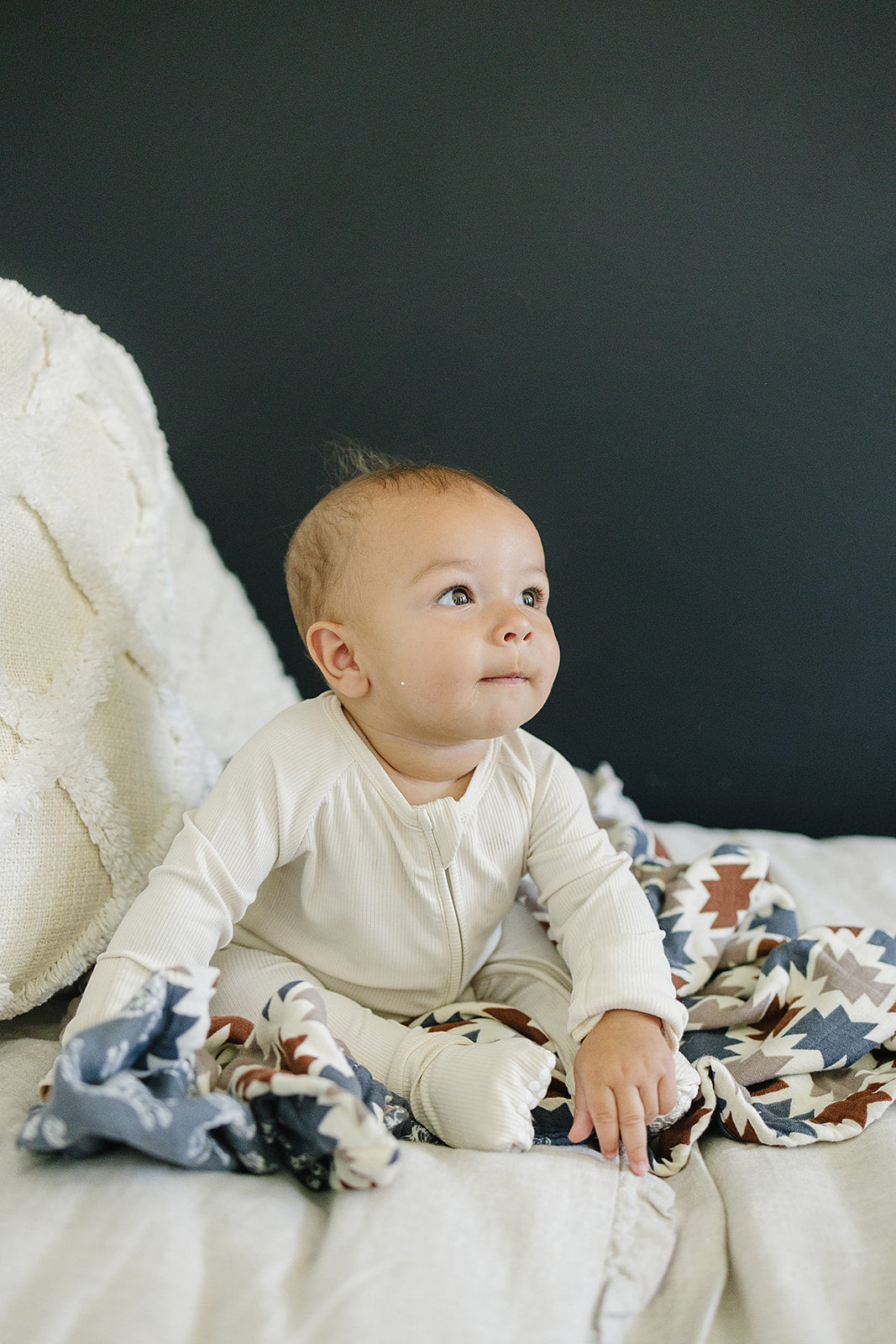 Baby wearing cream ribbed bamboo zipper footie, sitting on a soft bed with blankets, smiling adorably.