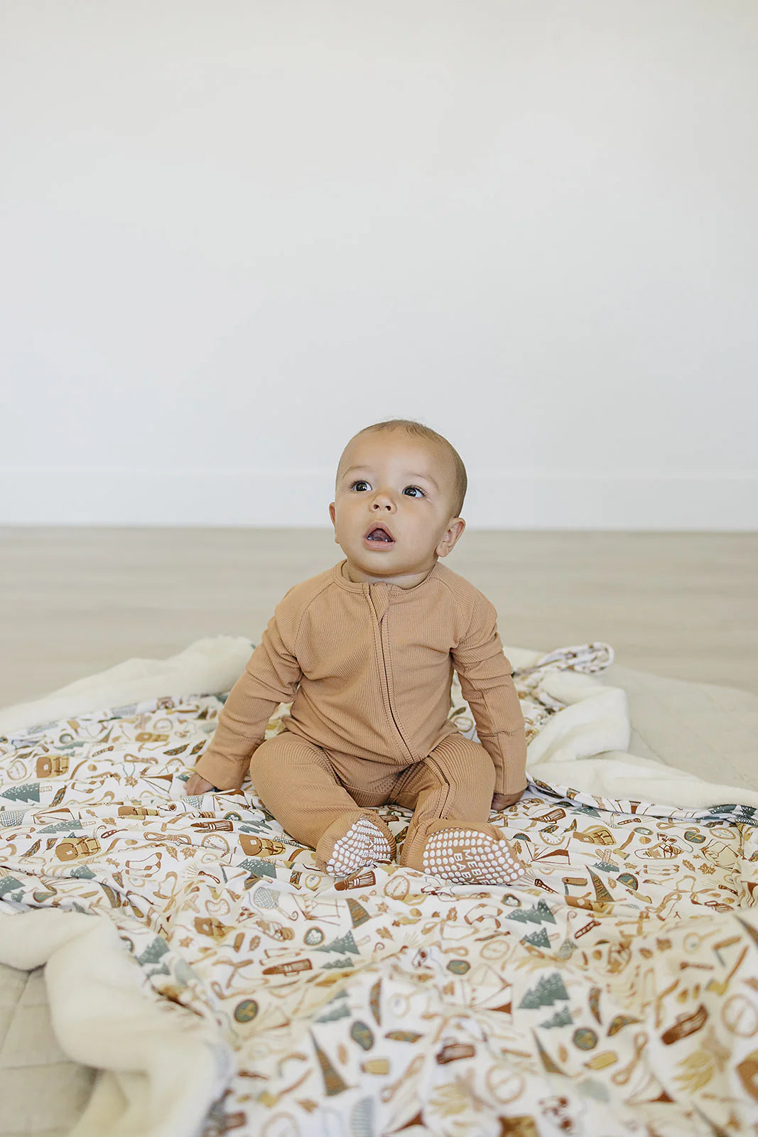 Baby in latte ribbed bamboo zipper footie sitting on a cozy patterned blanket, showcasing comfort and style.