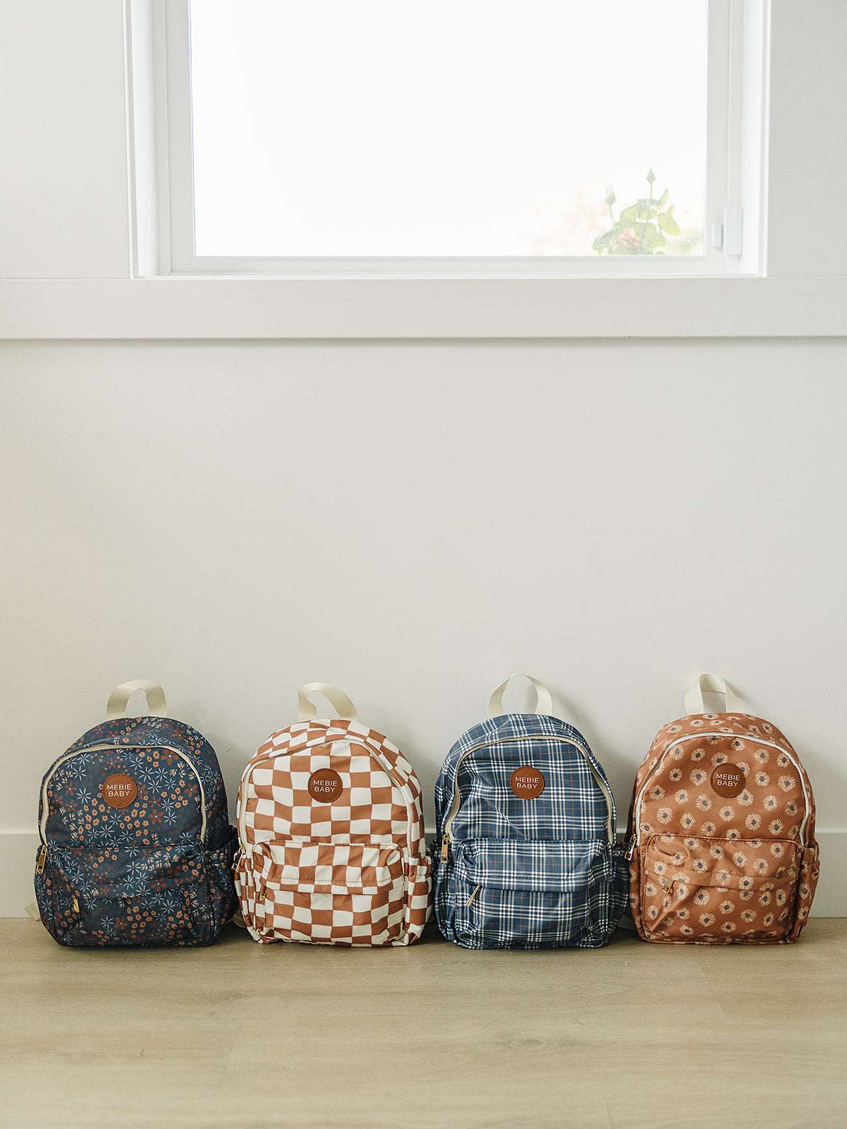Four stylish kids mini backpacks lined up against a wall, featuring various playful designs and colors.