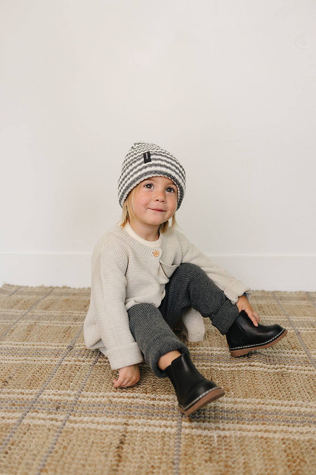 Baby wearing a grey and white baby beanie, cozy outfit, sitting on a woven rug.