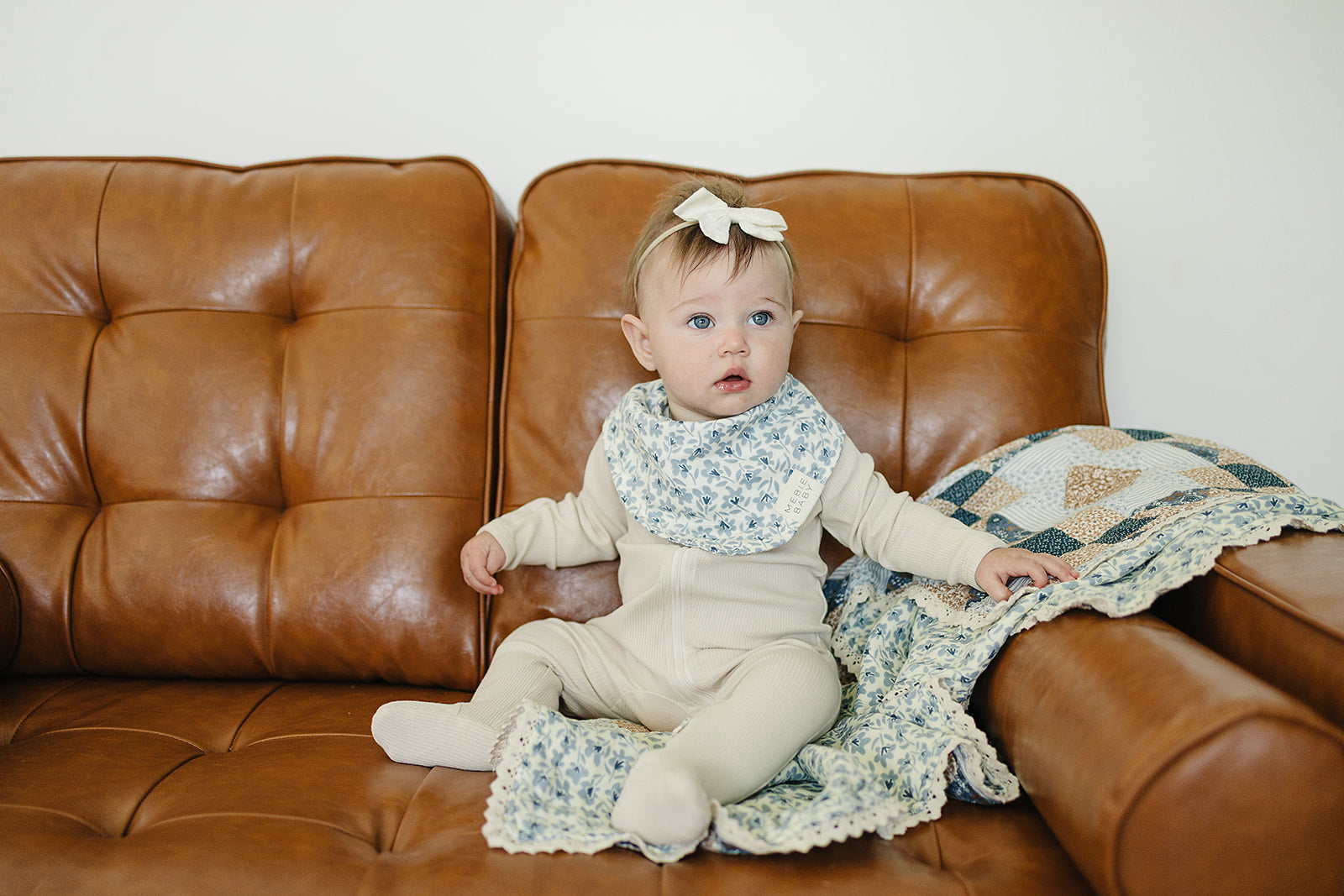 Baby wearing Blue Blossom Classic Muslin Bib by Mebie Baby, sitting on a brown couch with a colorful quilt.