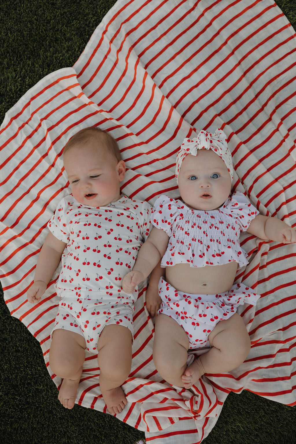 Two babies in strawberry print bamboo short sets on a striped blanket, perfect for summer playtime.
