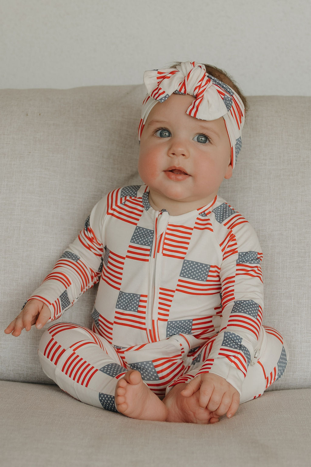 Baby in Americana bamboo zip pajamas with stars and stripes pattern, wearing a matching headband, sitting on a couch.