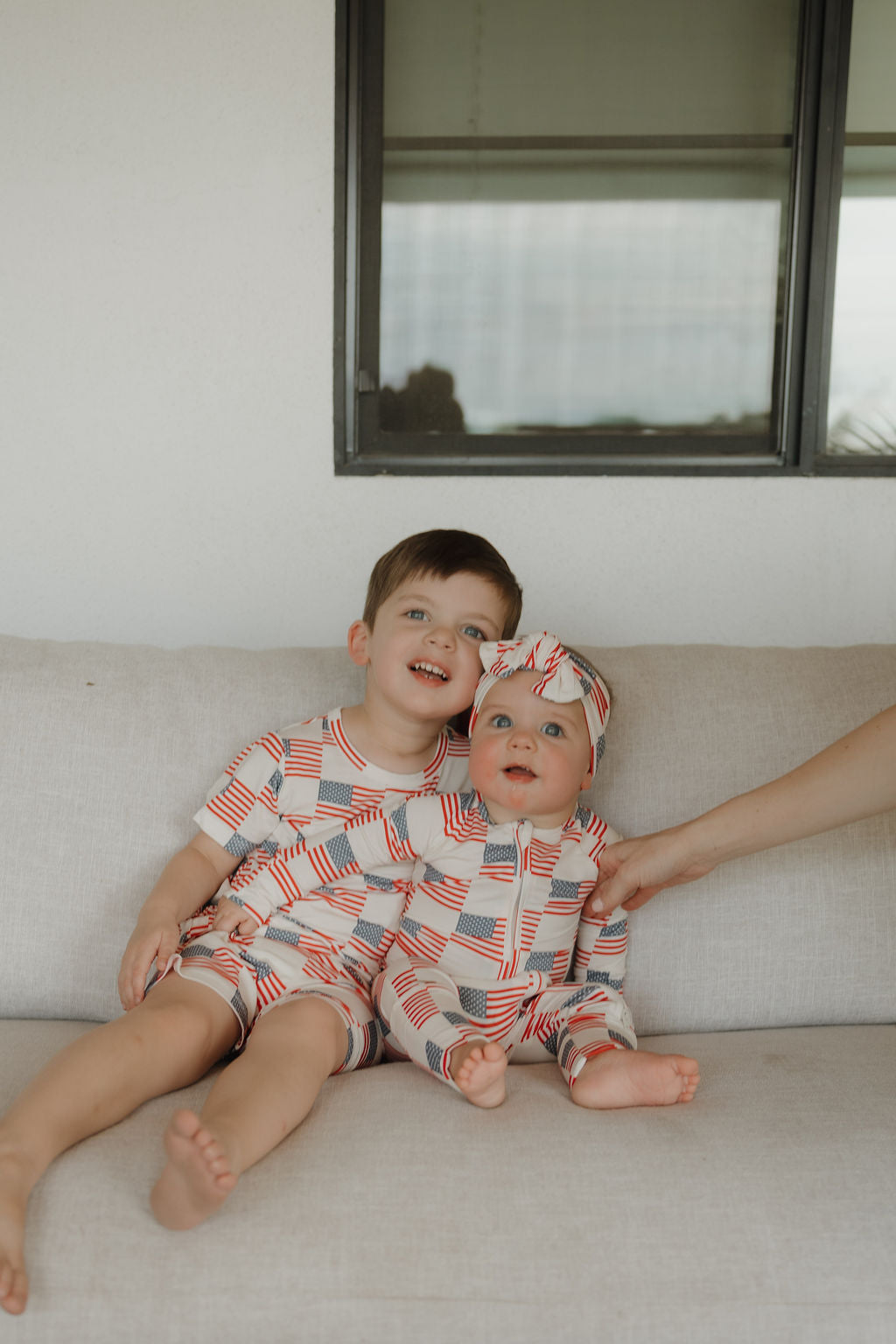 Two children in Americana-themed bamboo zip pajamas with stars and stripes patterns, sitting together on a sofa.