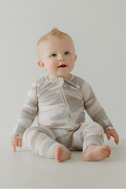 Cute baby in full-length bamboo zip pajamas with a neutral camo print, seated on a white background.