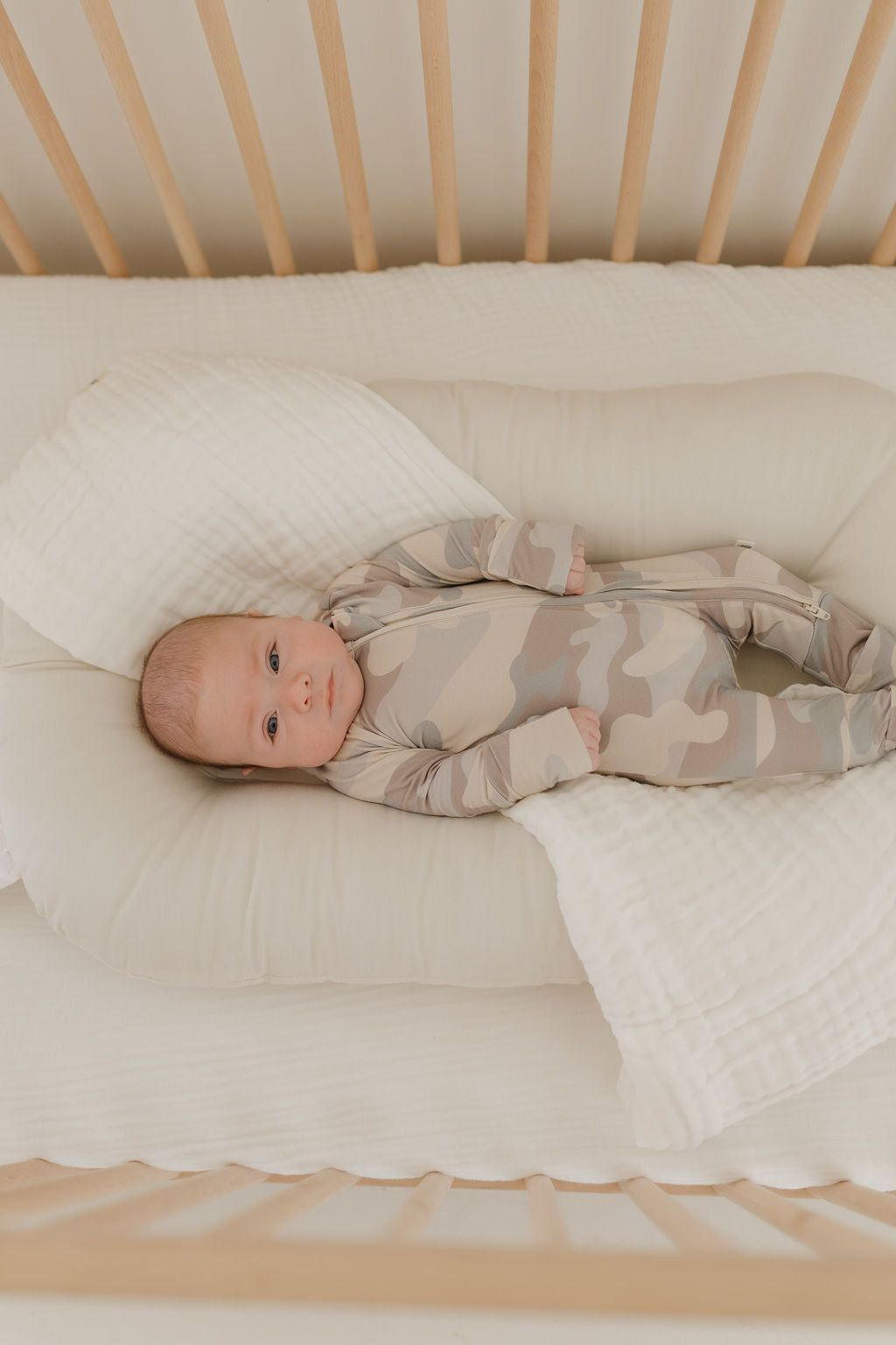Baby in neutral camo Bamboo Zip Pajamas, lying comfortably in a crib, showcasing stylish and cozy loungewear.