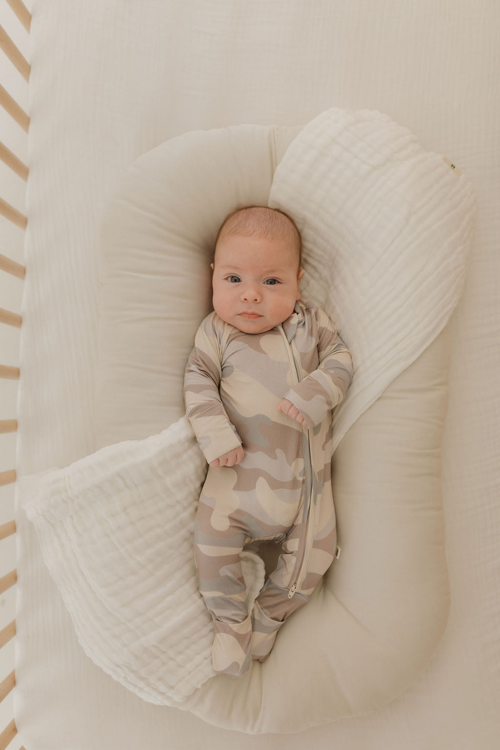 Baby in camouflage bamboo zip pajamas, nestled comfortably in a soft crib insert.