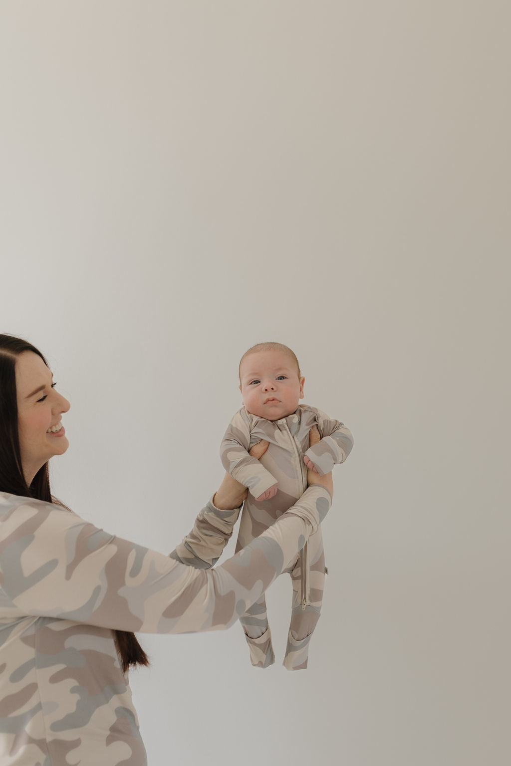 Mom holding a baby in bamboo zip pajamas with a neutral camo print against a plain background.