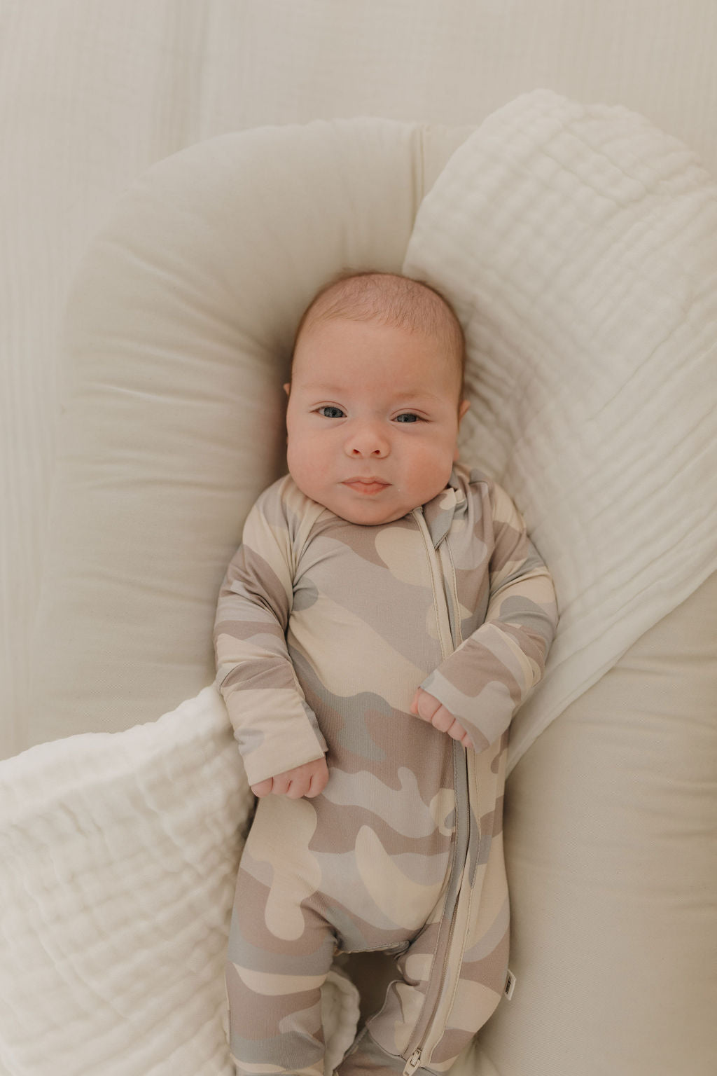 Baby in cozy bamboo zip pajamas with neutral camo print, lying on a soft surface.
