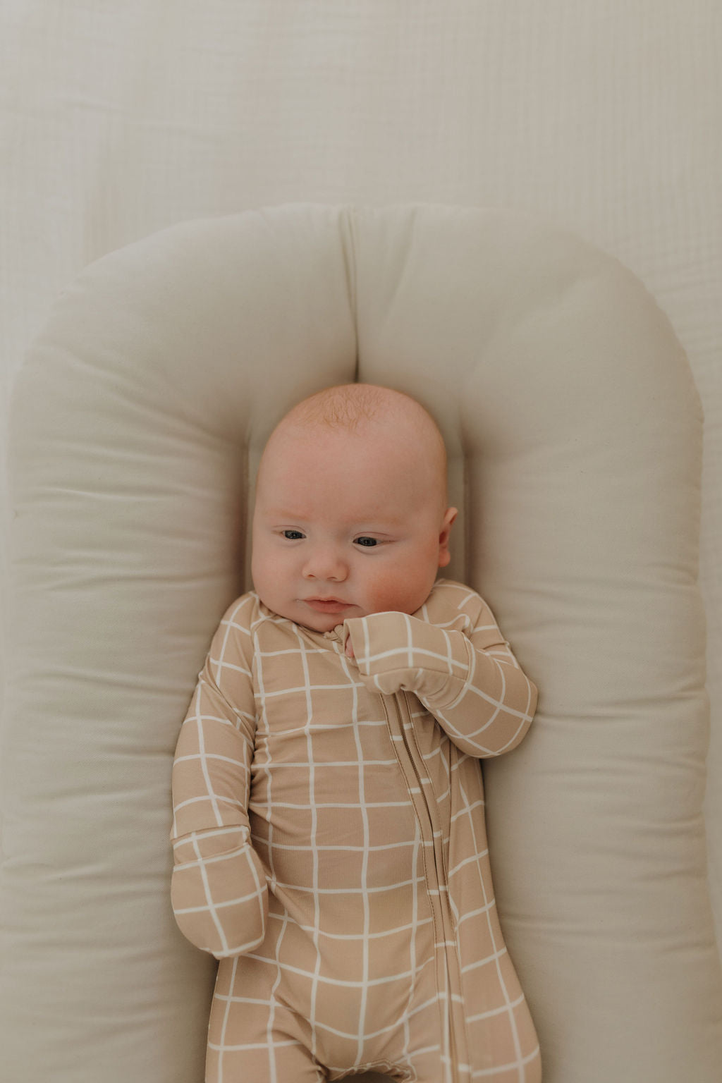 Baby wearing beige grid bamboo zip pajamas, resting comfortably in a baby nest with a calm expression.