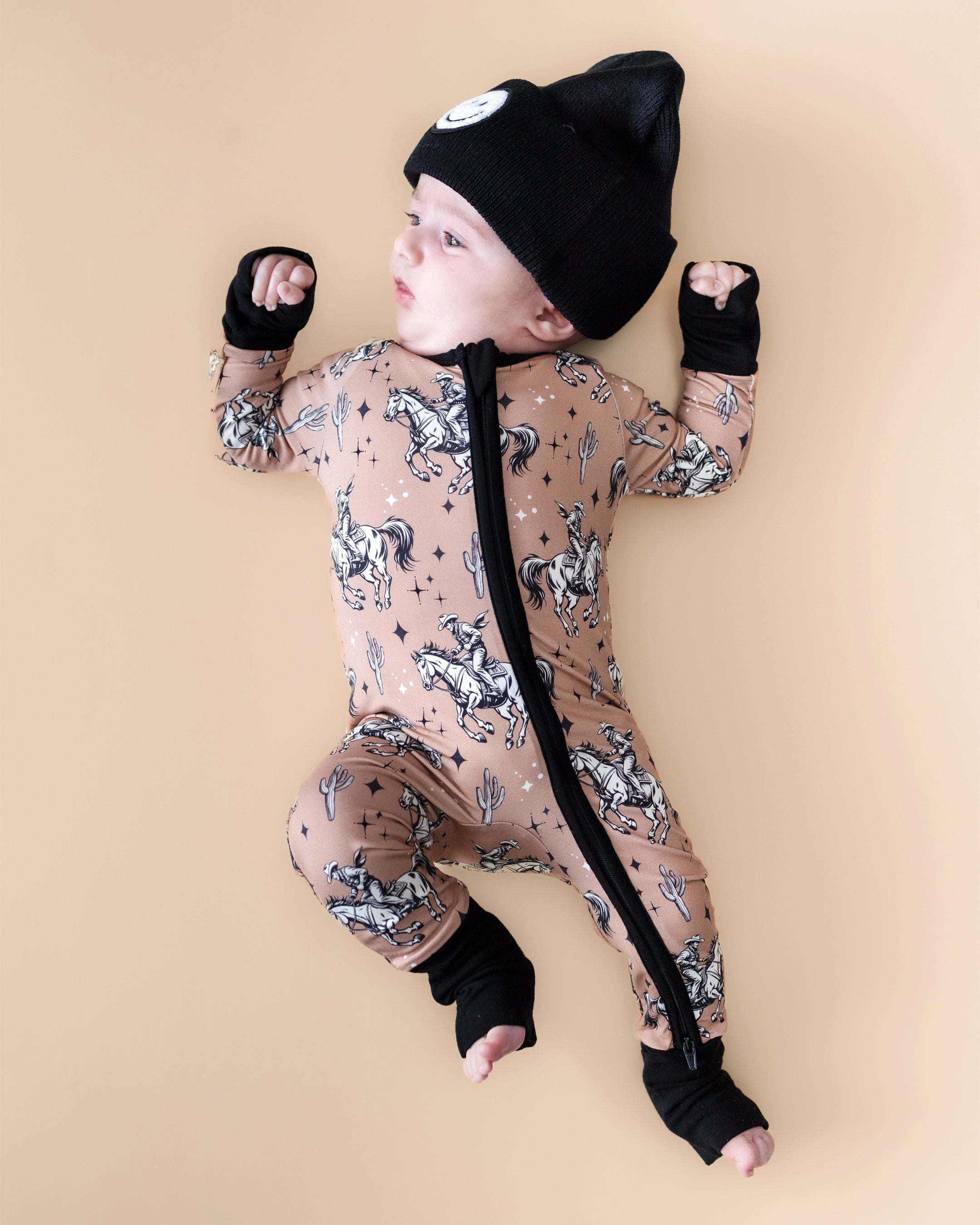 Baby in Bamboo Zip Romper with cowboy print and beanie hat, lying on a beige surface.