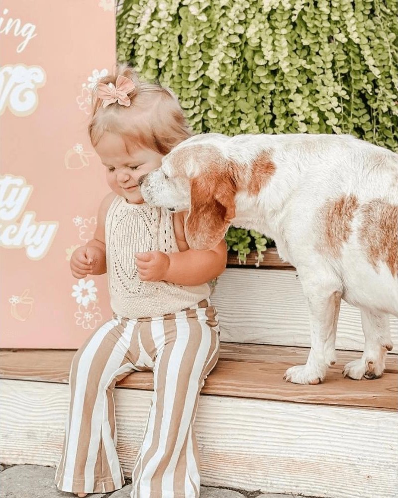Child in tan striped bell bottom pants, smiling with a dog, surrounded by greenery and a pink backdrop.