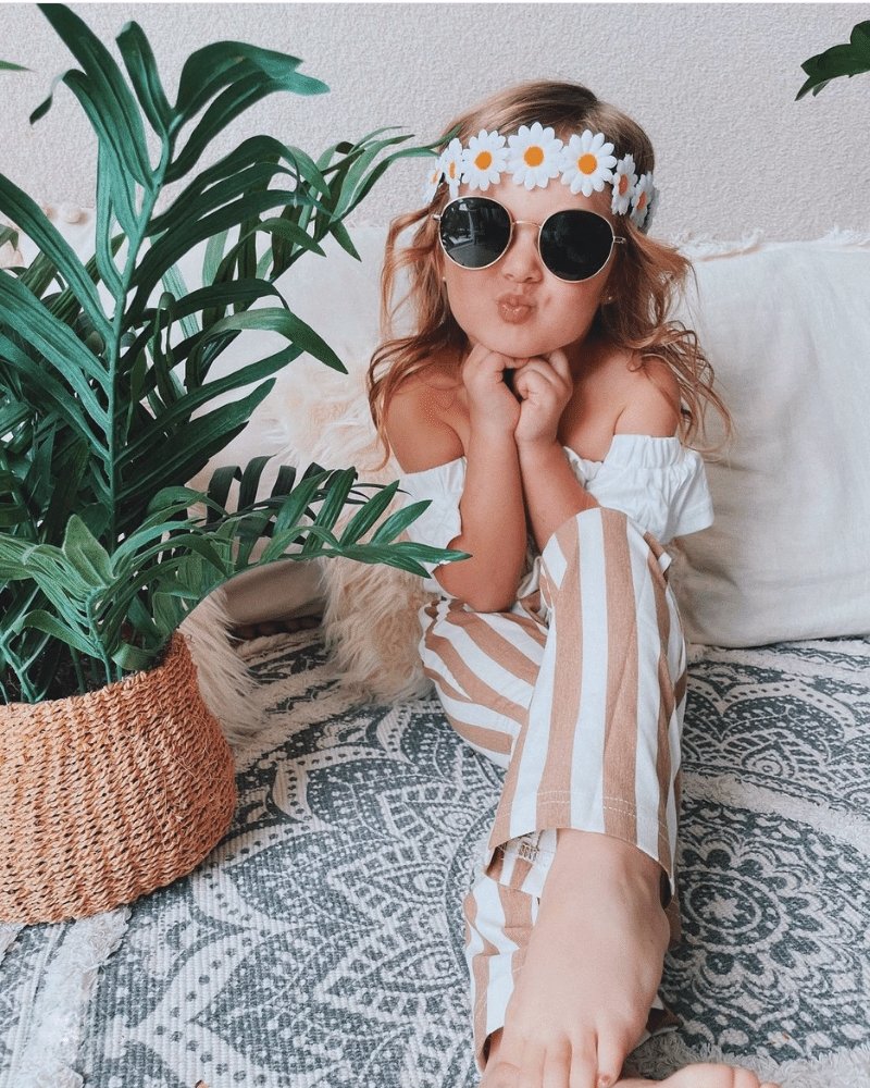 Kid wearing tan striped bell bottom pants, daisy flower crown, and sunglasses, posing playfully on a cozy blanket.