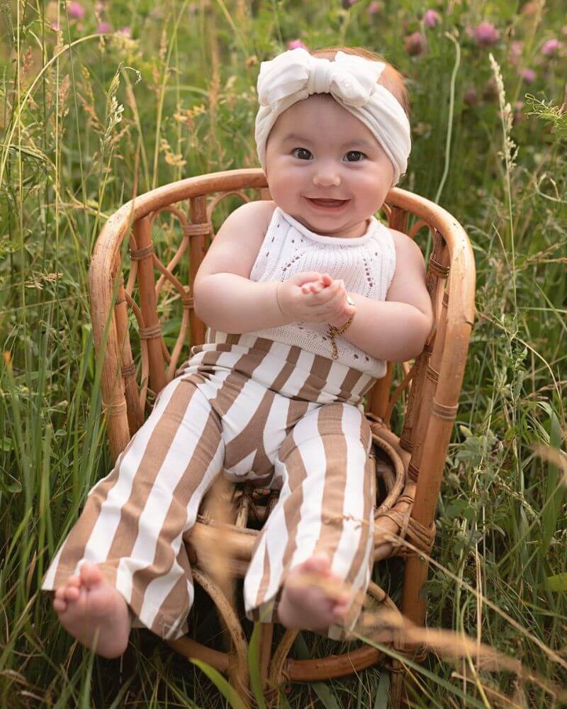 Smiling baby in tan striped bell bottom pants for kids, sitting in a chair surrounded by grass and flowers.