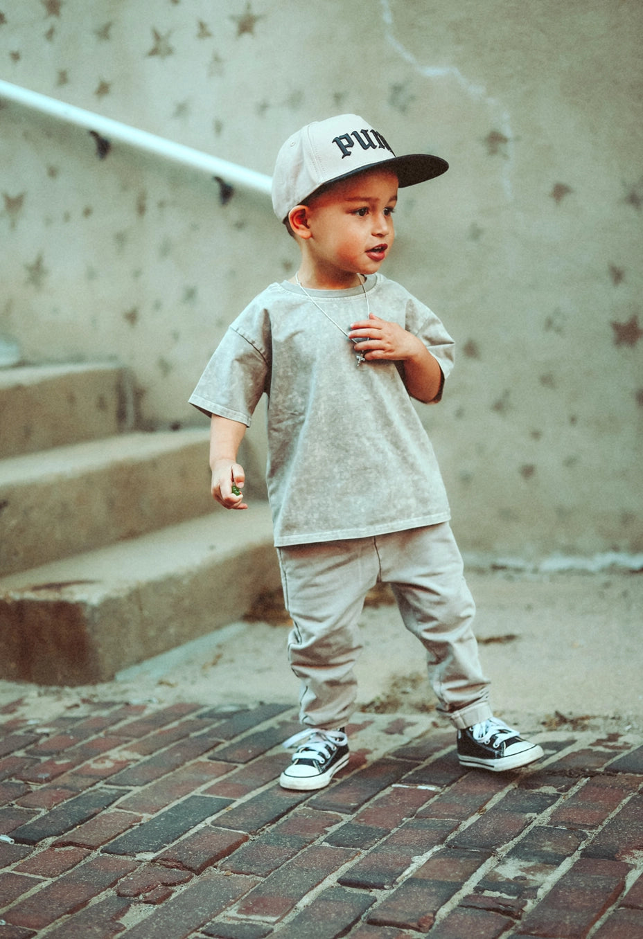 Toddler wearing tan acid wash joggers and matching t-shirt, standing on brick steps with a playful pose.