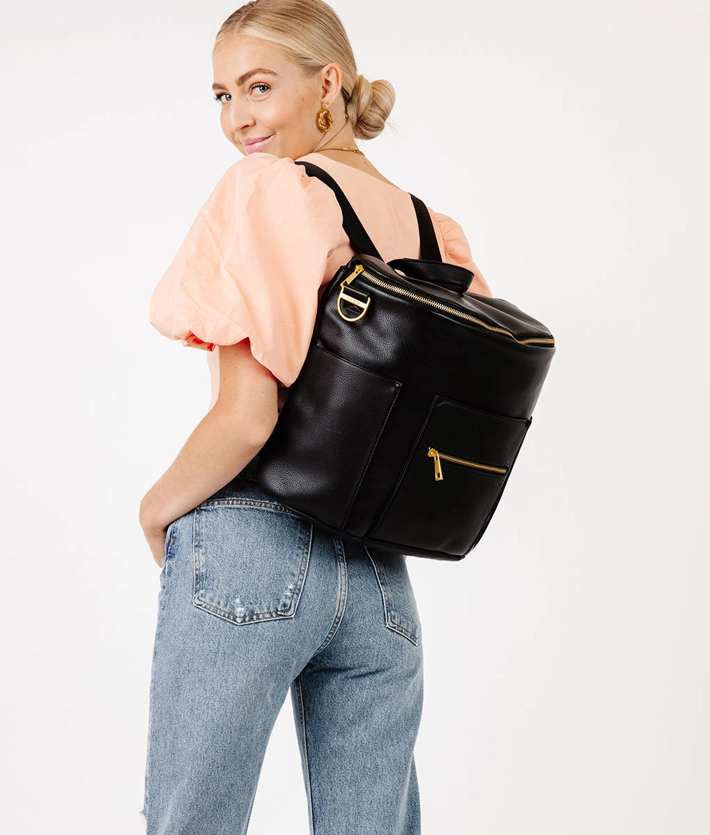 Stylish woman wearing the Original Diaper Bag Backpack in black faux leather with gold accents.