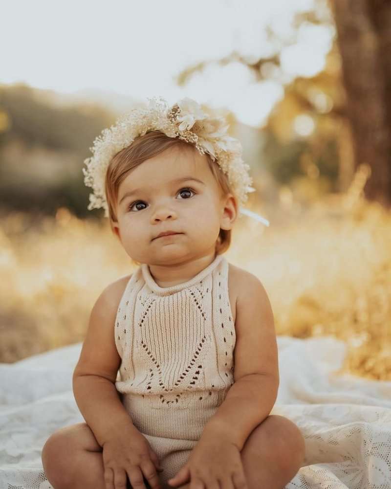 Baby wearing vanilla cream Drake crochet halter romper, sitting outdoors with floral headband and soft sunlight.